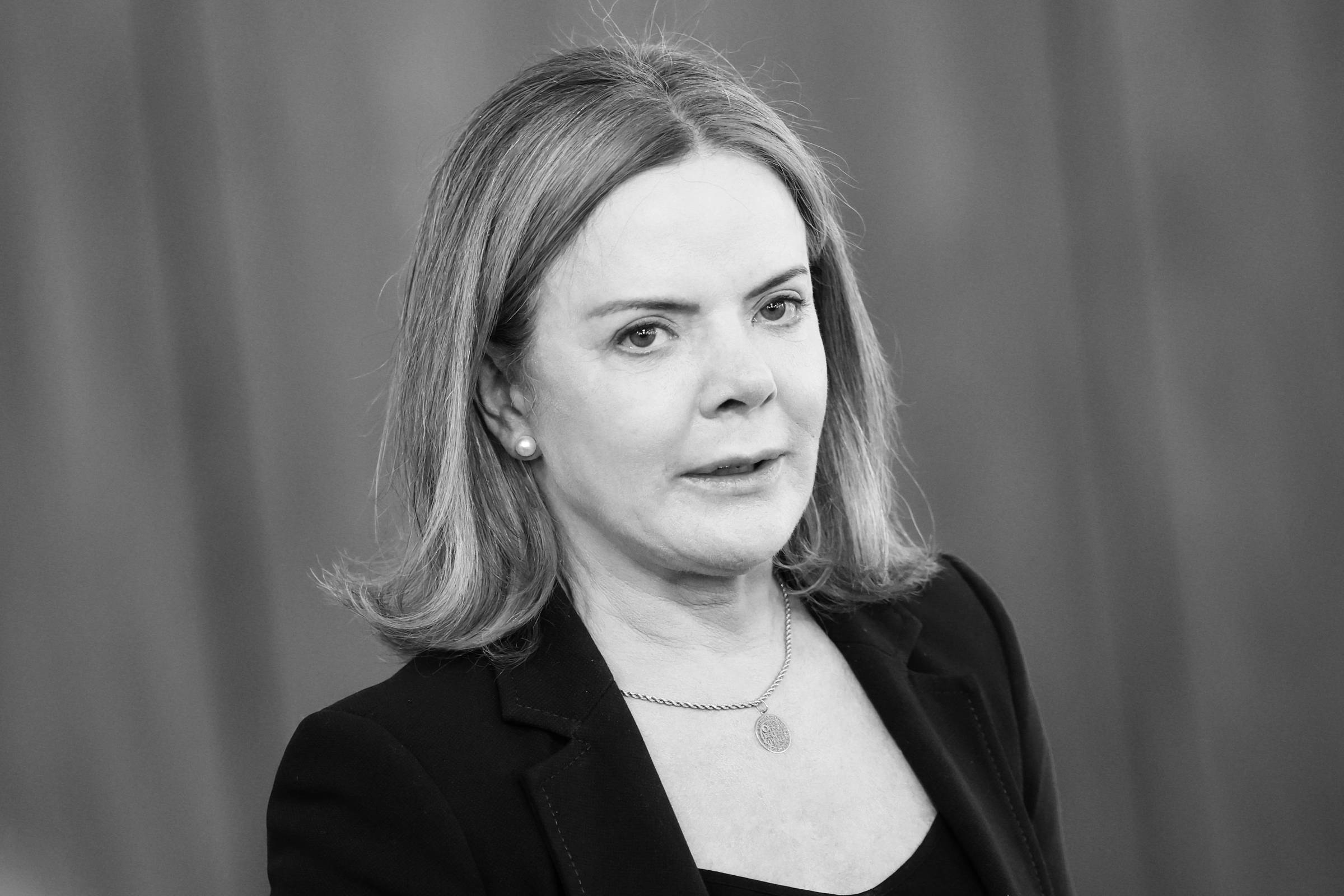 A imagem mostra uma mulher com cabelo liso e loiro, usando um blazer escuro e uma blusa preta. Ela está em um ambiente com fundo de madeira clara, olhando para a câmera com uma expressão séria. A mulher usa brincos de pérola e um colar discreto.