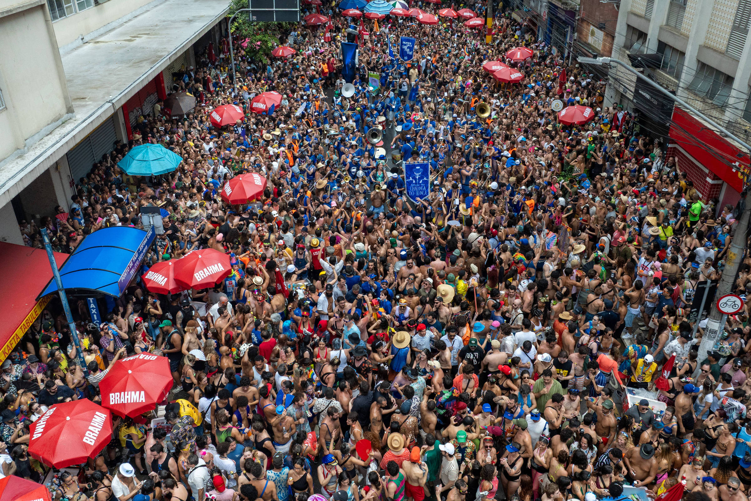A imagem mostra uma grande multidão reunida em uma rua, com pessoas vestindo roupas coloridas e algumas sem camisa. Há várias mesas e guarda-sóis vermelhos com a marca