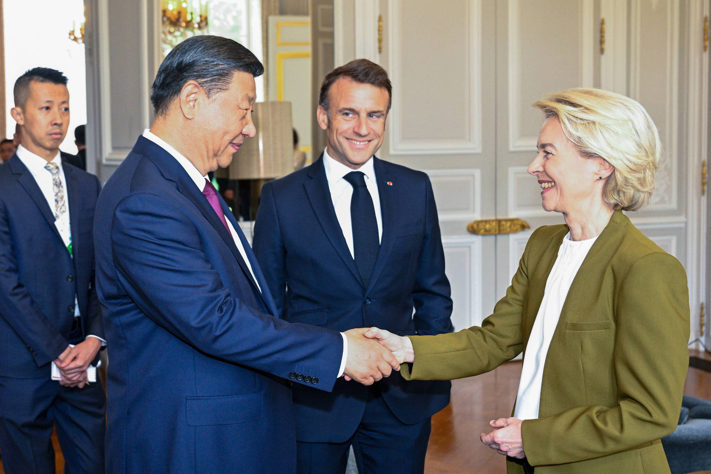 A imagem mostra um encontro entre três pessoas em um ambiente interno elegante. À esquerda, Xi Jinping, de terno escuro, está apertando a mão de von der Leyen, uma mulher de cabelo loiro e curto, que usa um blazer verde. Ao fundo, Macron, um homem de terno escuro, observa com um sorriso. O ambiente é bem iluminado, com paredes claras e detalhes decorativos.