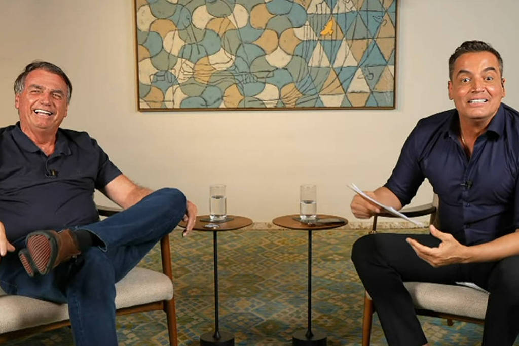 A imagem mostra duas pessoas sentadas em cadeiras confortáveis em um ambiente interno. À esquerda, um homem sorridente, vestido com uma camiseta escura e calças, está relaxado com as pernas cruzadas. À direita, outro homem, também sorridente, usa uma camisa de manga longa escura e está segurando um papel, parecendo animado. Entre eles, há duas mesas pequenas com copos de água. Ao fundo, uma obra de arte com formas geométricas em tons de azul e amarelo.