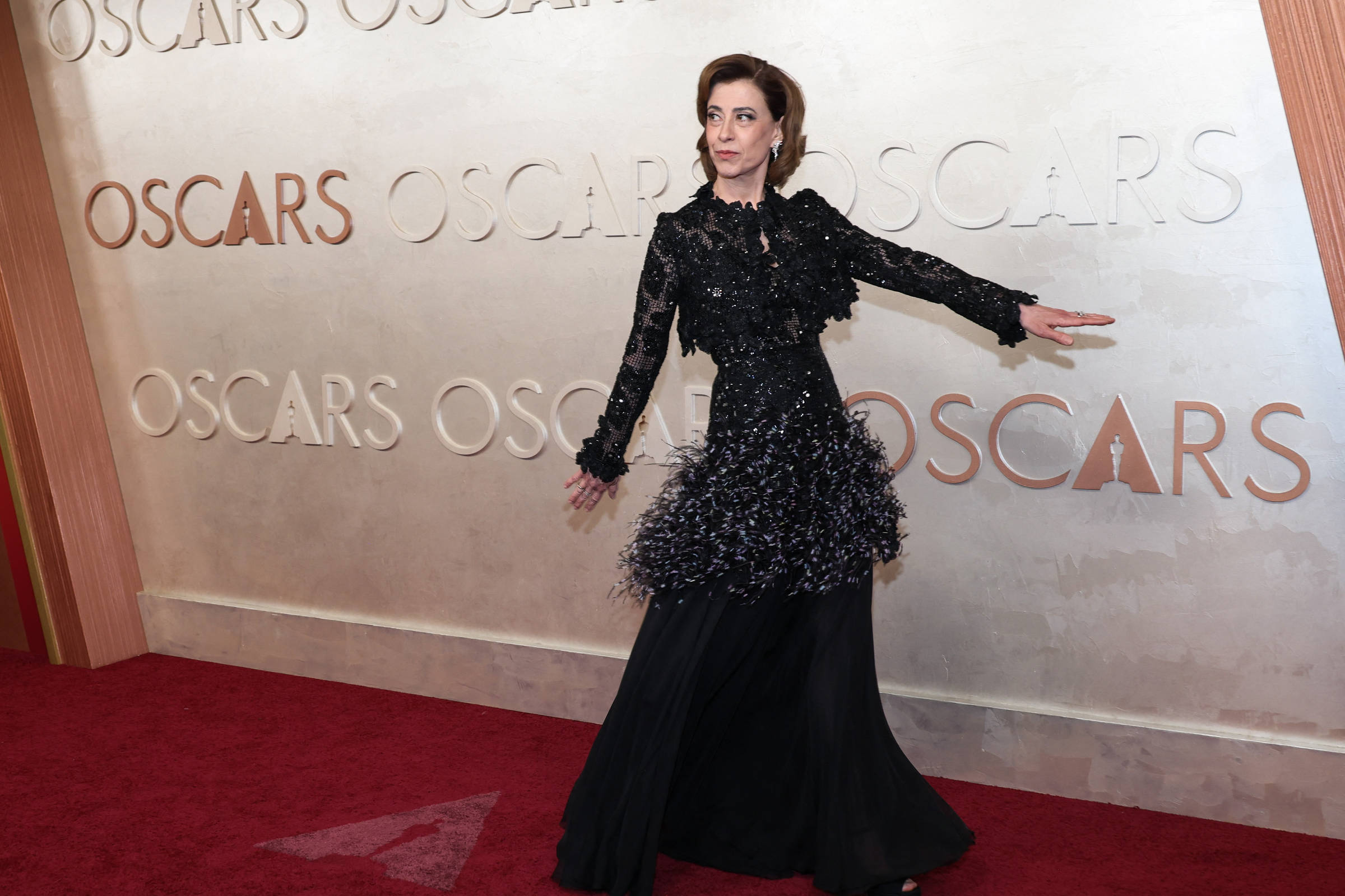 Uma mulher está posando em um tapete vermelho em um evento do Oscar. Ela usa um vestido preto com detalhes em brilho e uma saia volumosa. Ao fundo, há uma parede com a palavra
