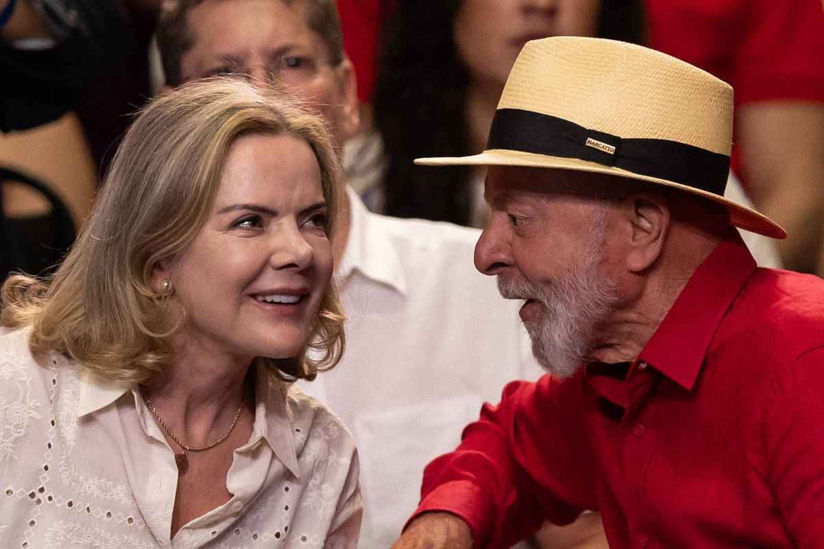 A imagem mostra duas pessoas conversando em um evento. À esquerda, uma mulher com cabelo loiro e ondulado, vestindo uma blusa clara com detalhes em renda. À direita, um homem idoso com barba branca, usando uma camisa vermelha e um chapéu de palha com uma faixa preta. Ao fundo, outras pessoas estão visíveis, algumas com roupas em tons de vermelho.