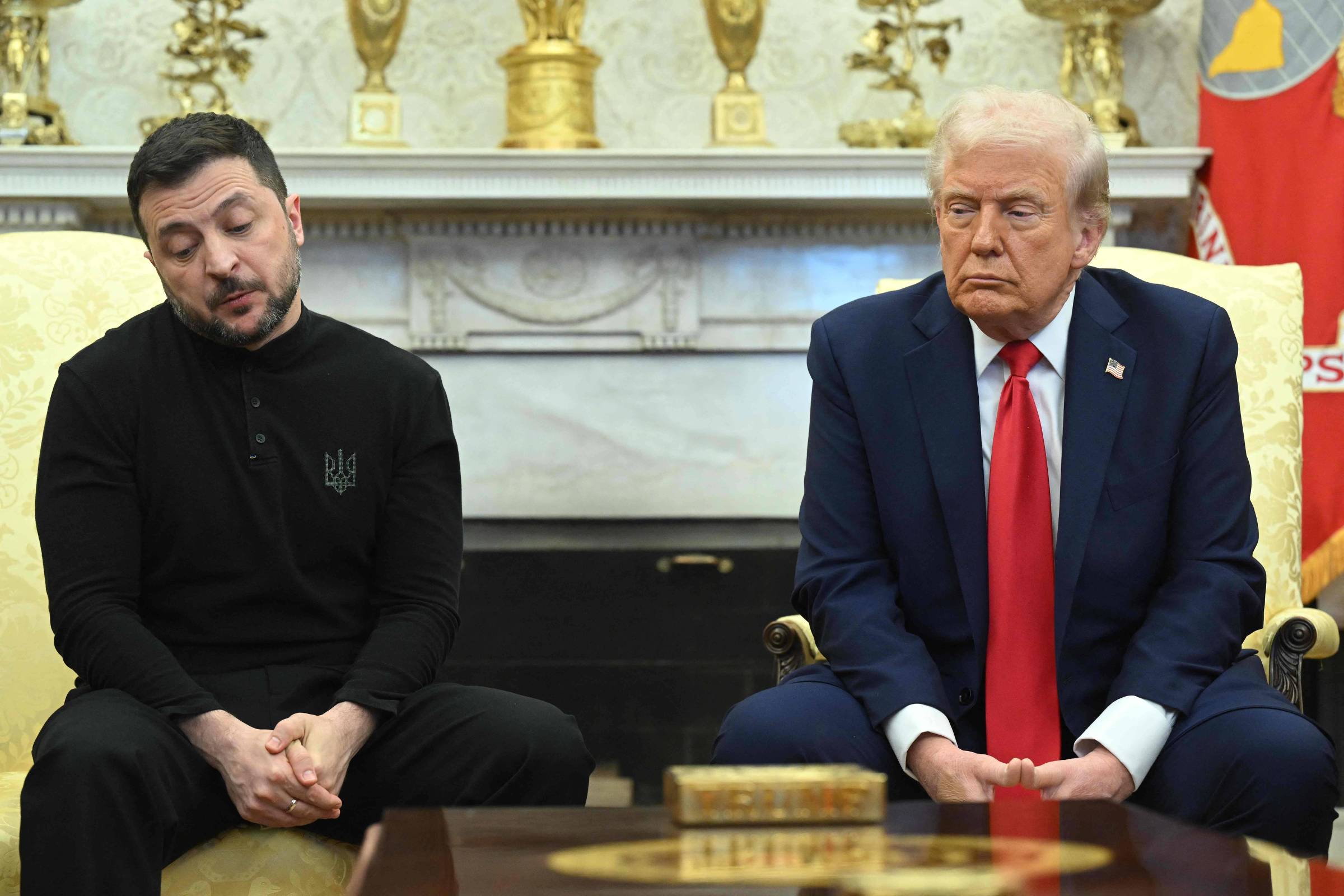 A imagem mostra duas pessoas sentadas em um sofá no Salão Oval da Casa Branca. À esquerda, Zelenski, um homem com cabelo escuro e barba, vestindo uma camiseta preta com o símbolo da Ucrânia. À direita, Trump, um homem com cabelo loiro e terno azul, com uma gravata vermelha, está sentado com uma expressão séria. Ao fundo, há uma lareira e prateleiras decoradas com objetos dourados.
