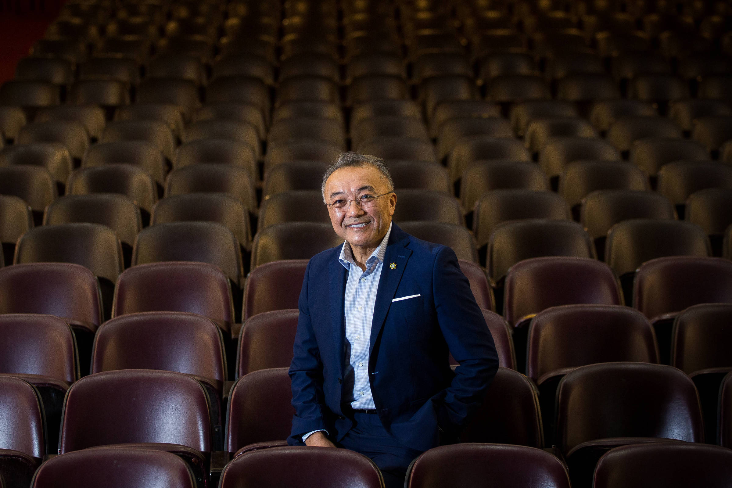 Um homem sorridente está posicionado em um auditório vazio, cercado por fileiras de cadeiras marrons. Ele usa um terno escuro e uma camisa clara, com um broche visível no paletó. O fundo mostra uma iluminação suave e as cadeiras se estendem em perspectiva, criando uma sensação de profundidade.