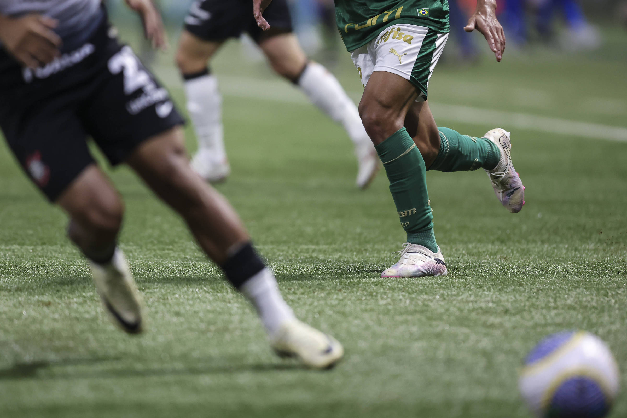 A imagem mostra jogadores de futebol em um campo, com foco nas pernas de um jogador vestindo um uniforme verde e branco, enquanto outro jogador, em uniforme preto, está em movimento ao fundo. A grama do campo é visível e a bola de futebol está parcialmente visível no canto inferior direito.