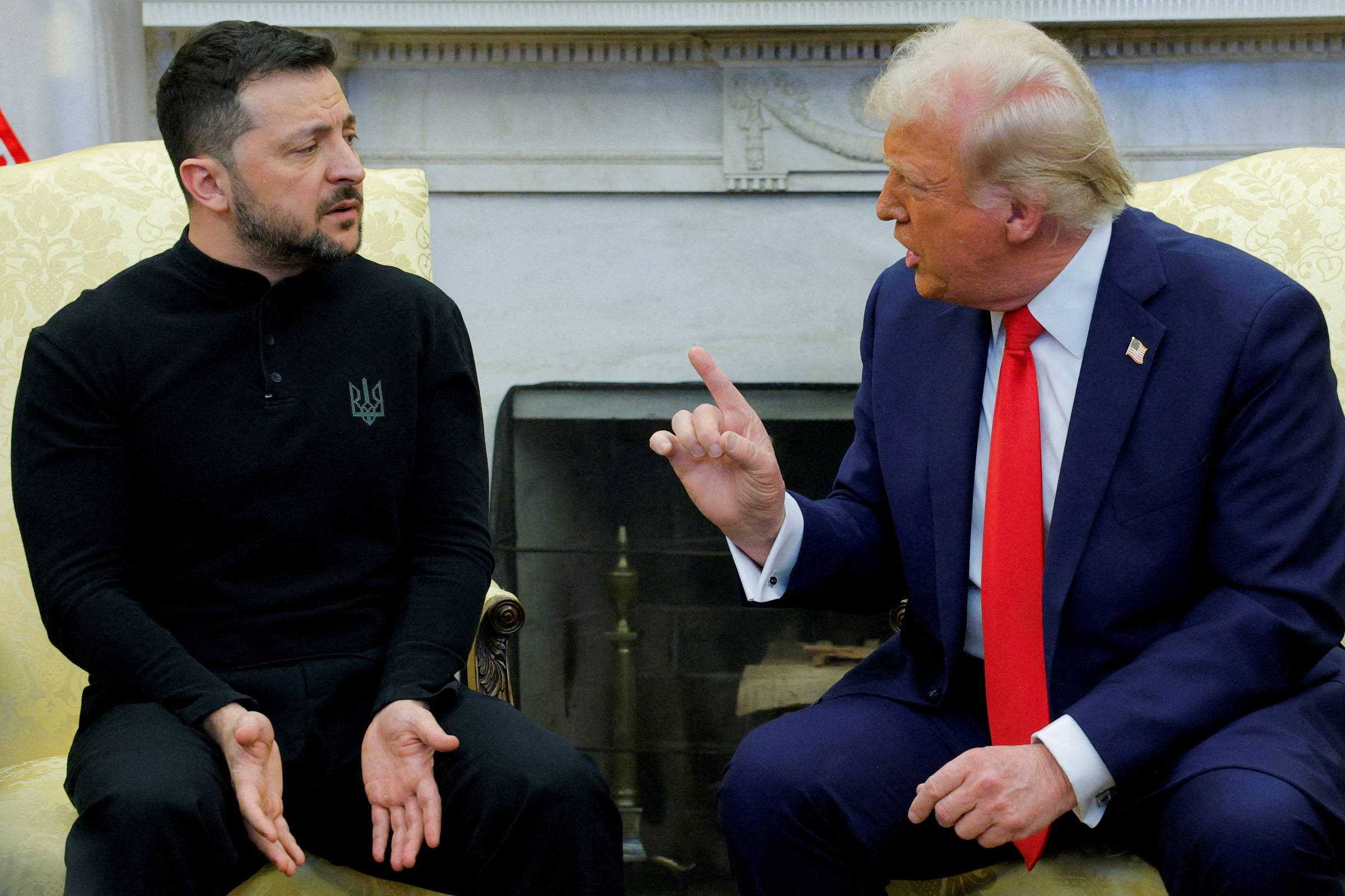 A imagem mostra dois homens sentados em um ambiente formal. À esquerda, um homem com cabelo escuro e barba, vestindo uma camisa preta com o símbolo da Ucrânia, parece estar ouvindo atentamente. À direita, um homem com cabelo loiro e terno azul, com uma gravata vermelha, está gesticulando com a mão direita, parecendo falar de forma enfática. Ao fundo, há uma lareira e uma decoração clássica.