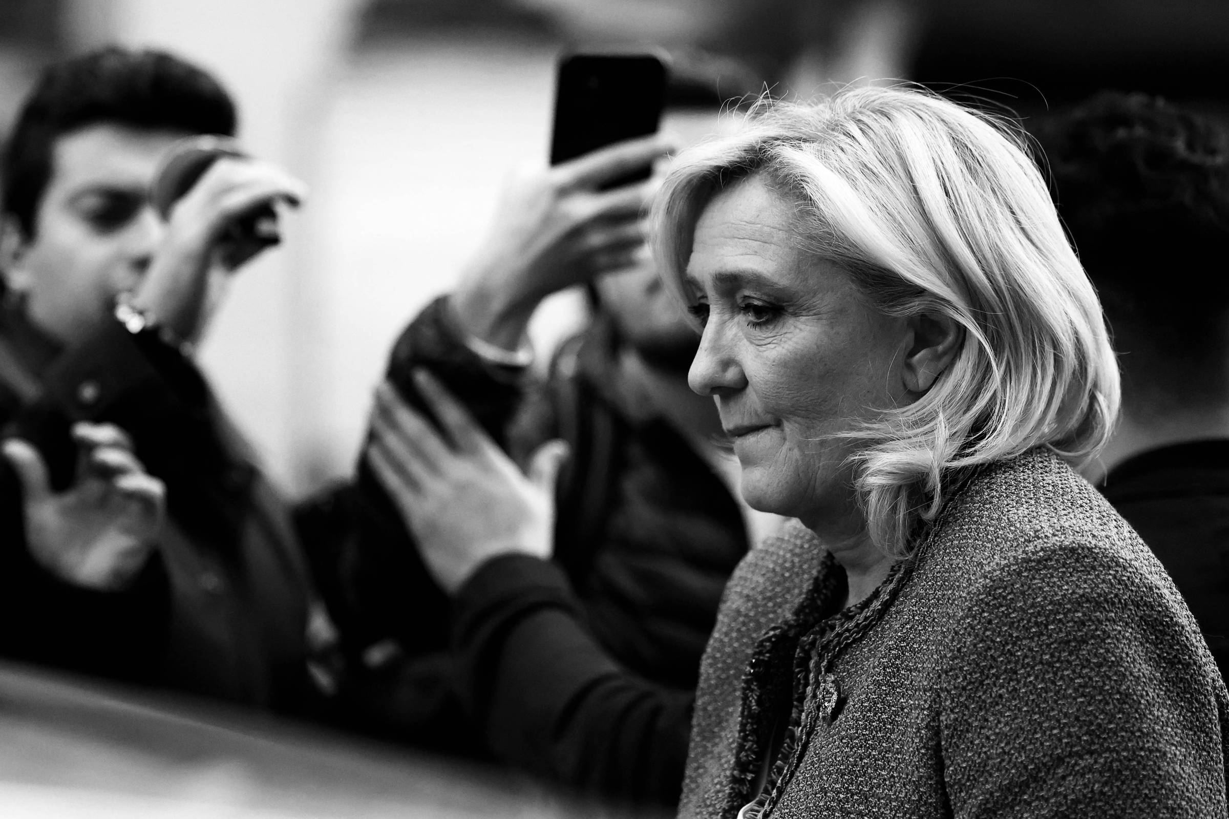 A imagem mostra uma mulher com cabelo loiro e liso, vestindo um casaco azul, caminhando em meio a um grupo de pessoas. Algumas pessoas ao fundo estão segurando celulares, aparentemente tirando fotos. O ambiente parece ser urbano, com uma atmosfera de agitação.