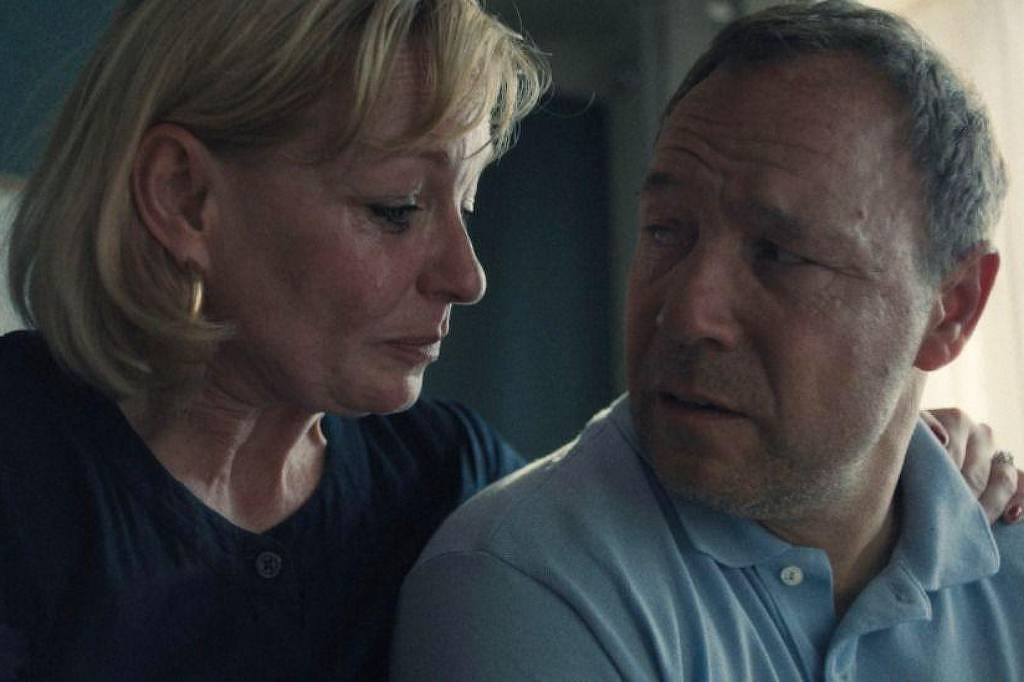 A imagem mostra duas pessoas em um momento íntimo e emocional. Uma mulher com cabelo loiro e curto, usando uma blusa escura, está olhando para um homem com cabelo curto e barba rala, que veste uma camisa polo clara. Eles parecem estar em uma conversa profunda, com expressões faciais que transmitem preocupação e empatia. O fundo é desfocado, sugerindo um ambiente acolhedor.