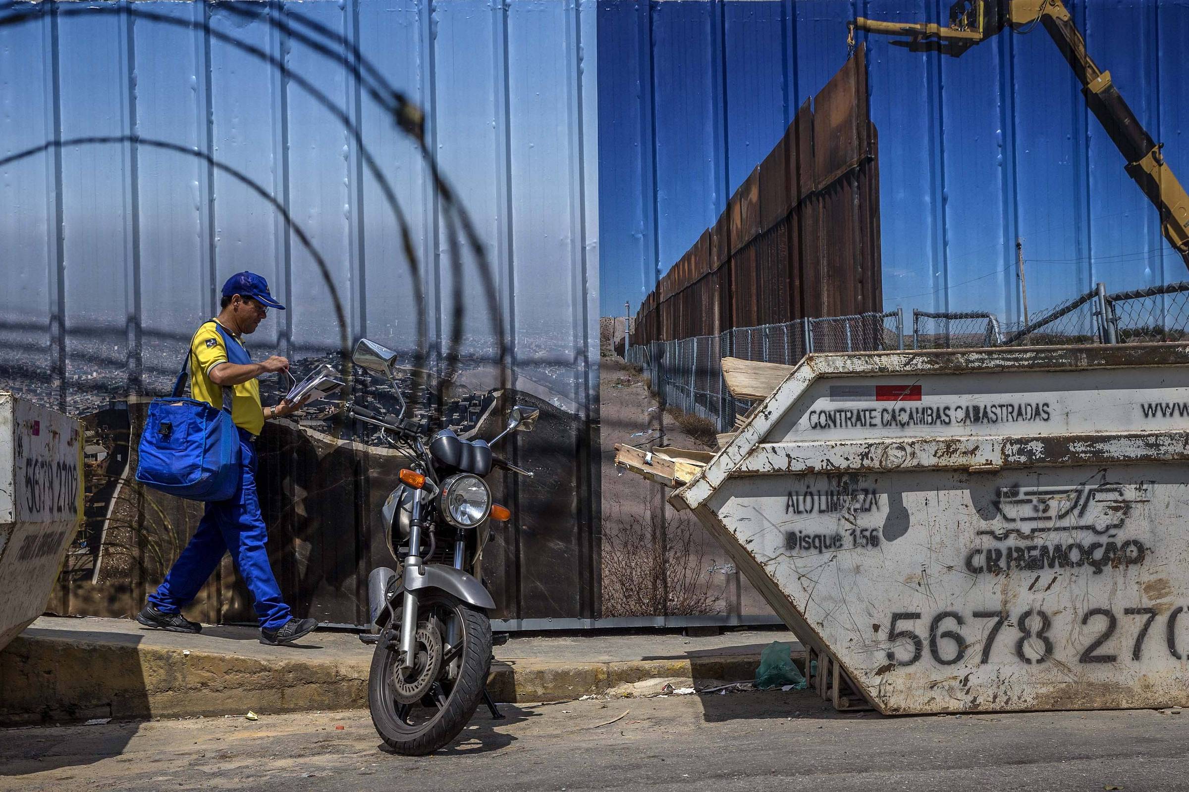 A imagem mostra um trabalhador vestido com uma camisa amarela e azul, carregando uma bolsa azul, caminhando em direção a uma motocicleta estacionada. Ao fundo, há um painel de construção com uma imagem de uma cerca e um guindaste. Dois contêineres de lixo estão visíveis na parte inferior da imagem, com inscrições e números.