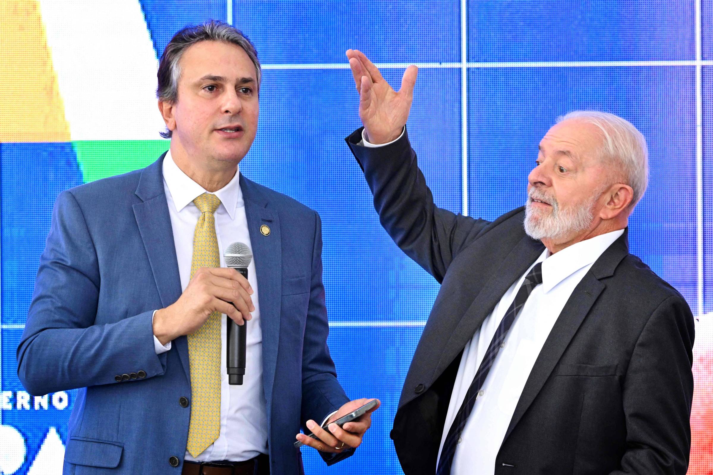 Homem alto de terno e gravata segura microfone. Ao seu lado, outro homem, de terno e gravata também, gesticula em sinal de deferência. O fundo é azul e parece indicar um telão