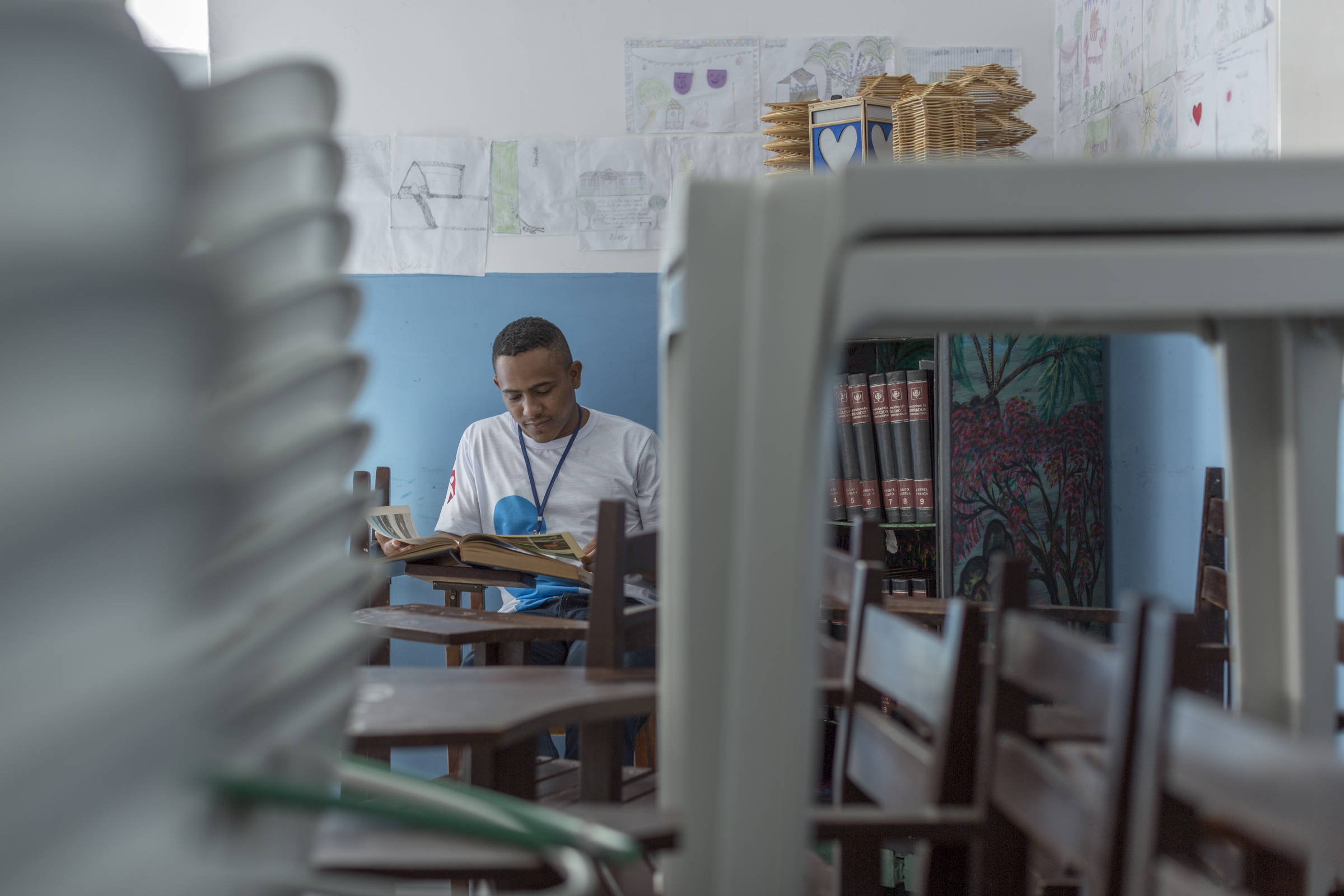Um jovem sentado em uma sala de aula, lendo um livro. O ambiente é decorado com desenhos nas paredes e há estantes com livros ao fundo. Cadeiras e mesas estão visíveis em primeiro plano, algumas empilhadas, sugerindo que a sala não está em uso no momento.
