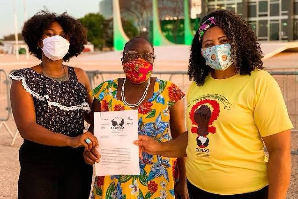 Três mulheres estão em pé, usando máscaras, em um ambiente externo. A mulher à esquerda veste uma blusa preta com detalhes brancos e calças escuras. A mulher do meio, que usa um vestido colorido, segura um documento. A mulher à direita está vestindo uma camiseta amarela com uma estampa e calças escuras. Ao fundo, há uma estrutura moderna e cercas de proteção.