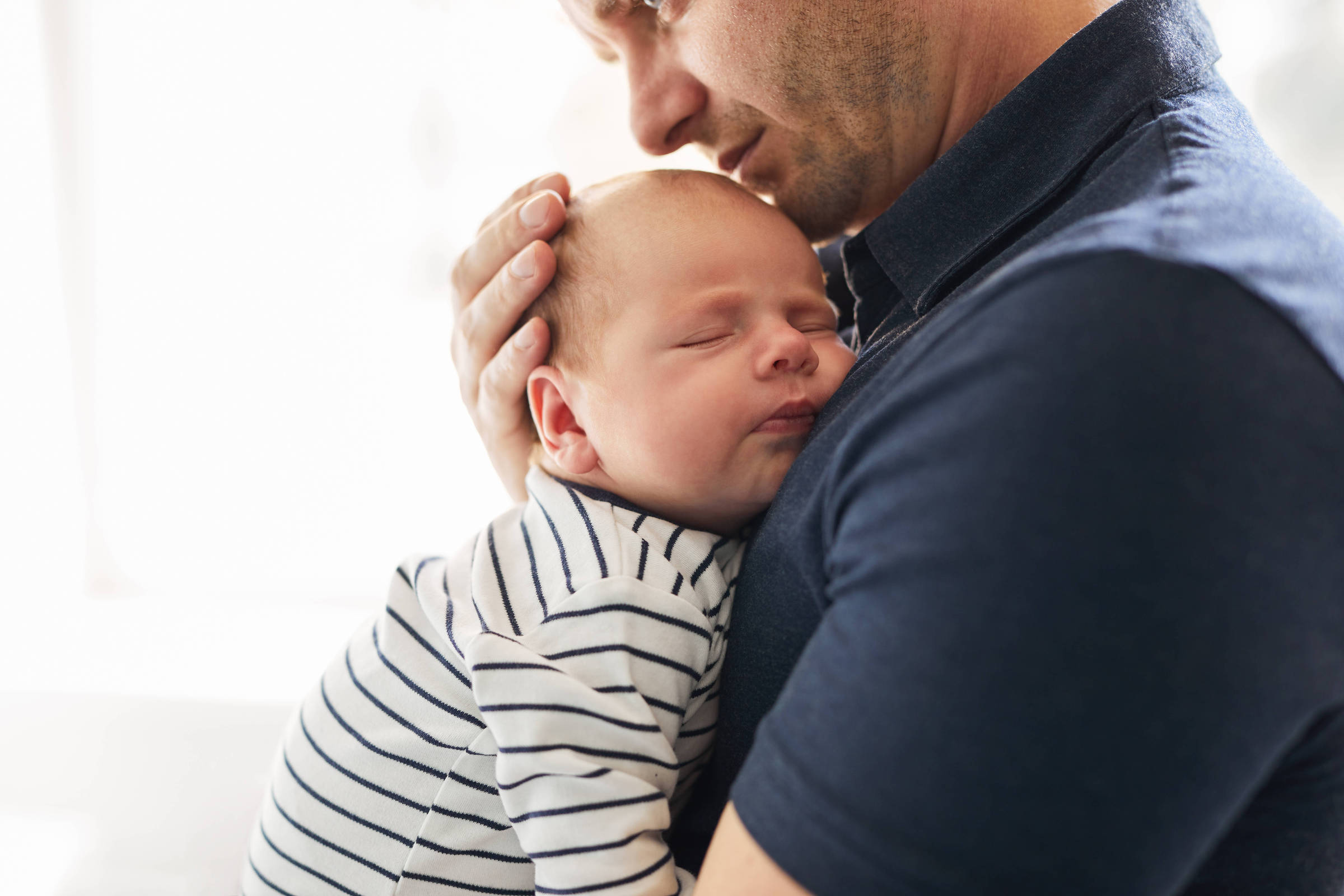 Um homem está segurando um bebê adormecido em seus braços. O bebê está vestido com uma roupa listrada em branco e azul, enquanto o homem usa uma camisa escura. O ambiente é iluminado, sugerindo um espaço acolhedor e tranquilo.
