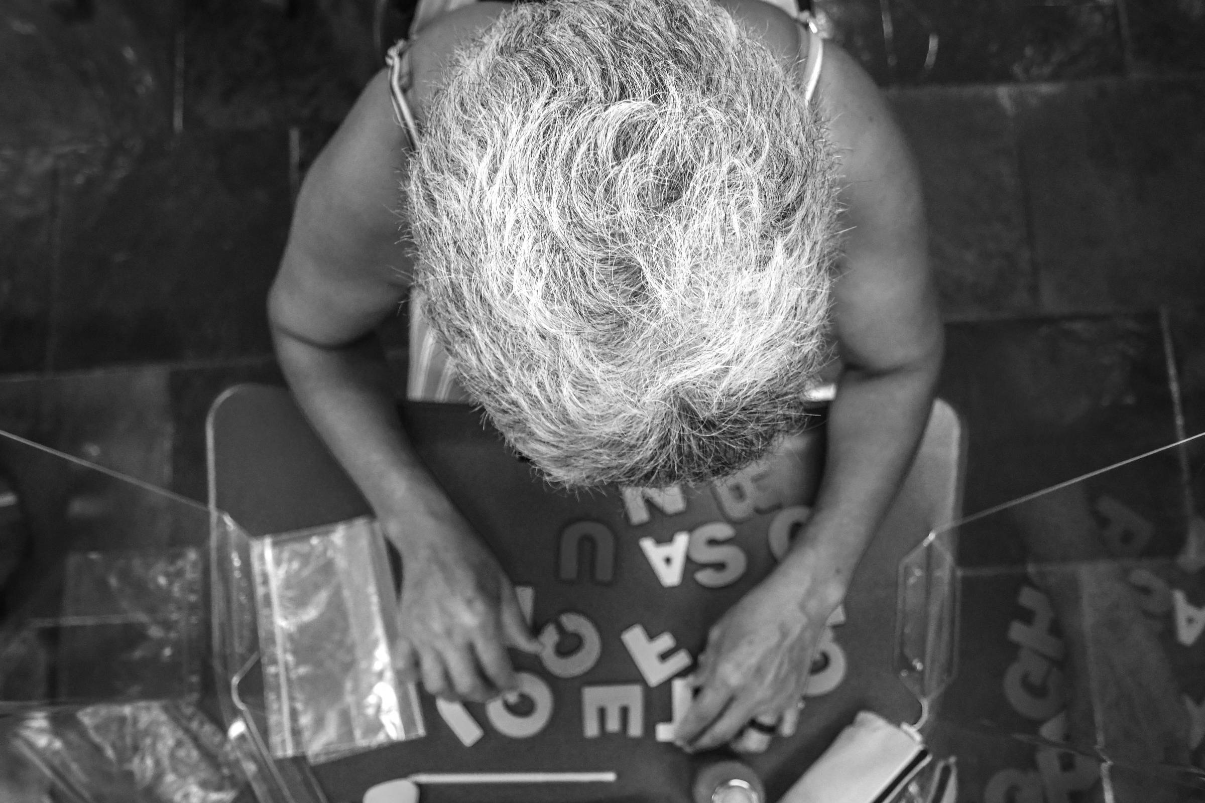 A imagem mostra a parte superior da cabeça de uma pessoa idosa com cabelo grisalho, que está sentada em uma mesa. A pessoa está manipulando letras coloridas de espuma dispostas em uma superfície azul. Ao lado, há um objeto rosa e um recipiente metálico. O ambiente parece ser um espaço de atividades, com piso de pedra e algumas cadeiras ao fundo.