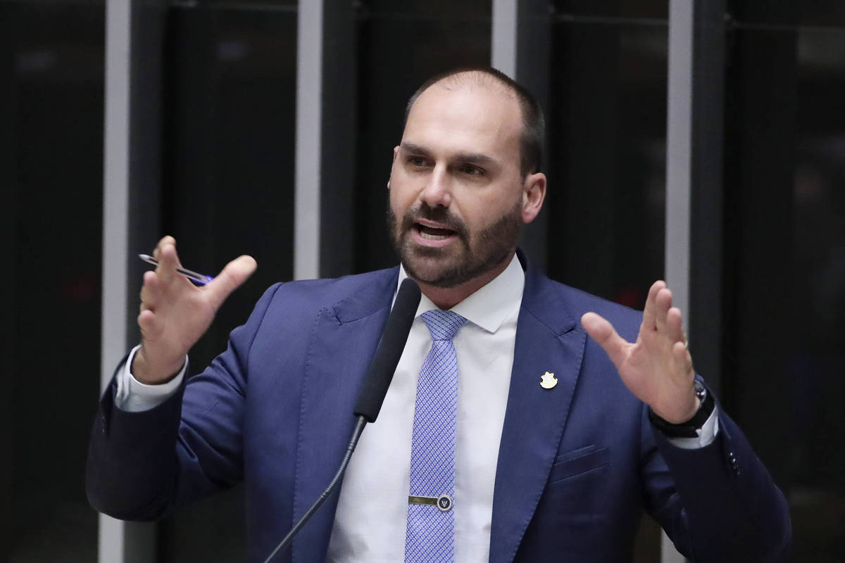 Um homem em um terno azul está falando em um microfone em um ambiente de plenário. Ele gesticula com as mãos, demonstrando entusiasmo ou ênfase em suas palavras. O fundo é composto por painéis de vidro, sugerindo um ambiente formal e institucional.