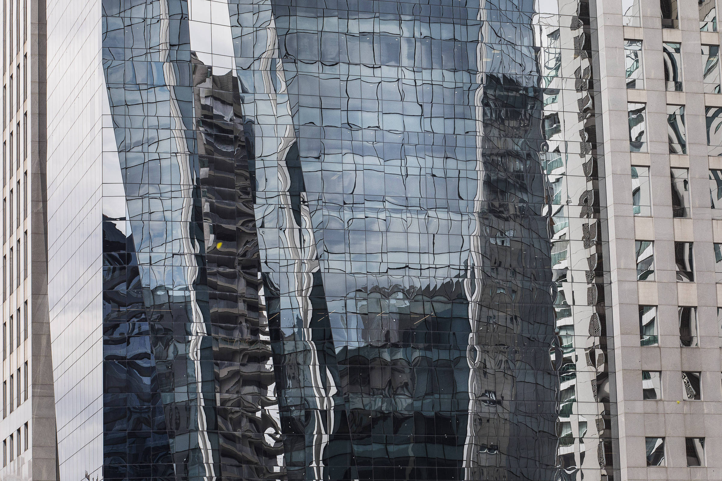 A imagem mostra a fachada de um edifício moderno com janelas de vidro refletindo outros prédios ao redor. A superfície do vidro apresenta um efeito distorcido, criando um padrão ondulado. À direita, há um edifício com uma fachada de concreto e janelas retangulares. Na parte inferior esquerda, uma árvore verde é visível, contrastando com a estrutura urbana.
