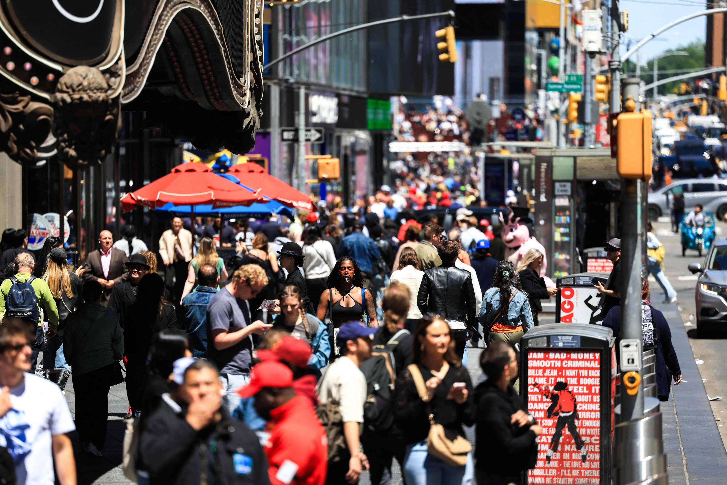 A imagem mostra uma rua movimentada em Times Square, Nova Iorque, com muitas pessoas caminhando. Há mesas com guarda-sóis vermelhos em um lado da calçada e várias lojas e anúncios visíveis ao fundo. O clima parece ensolarado e há uma grande quantidade de pessoas, incluindo turistas e locais, interagindo no espaço público.