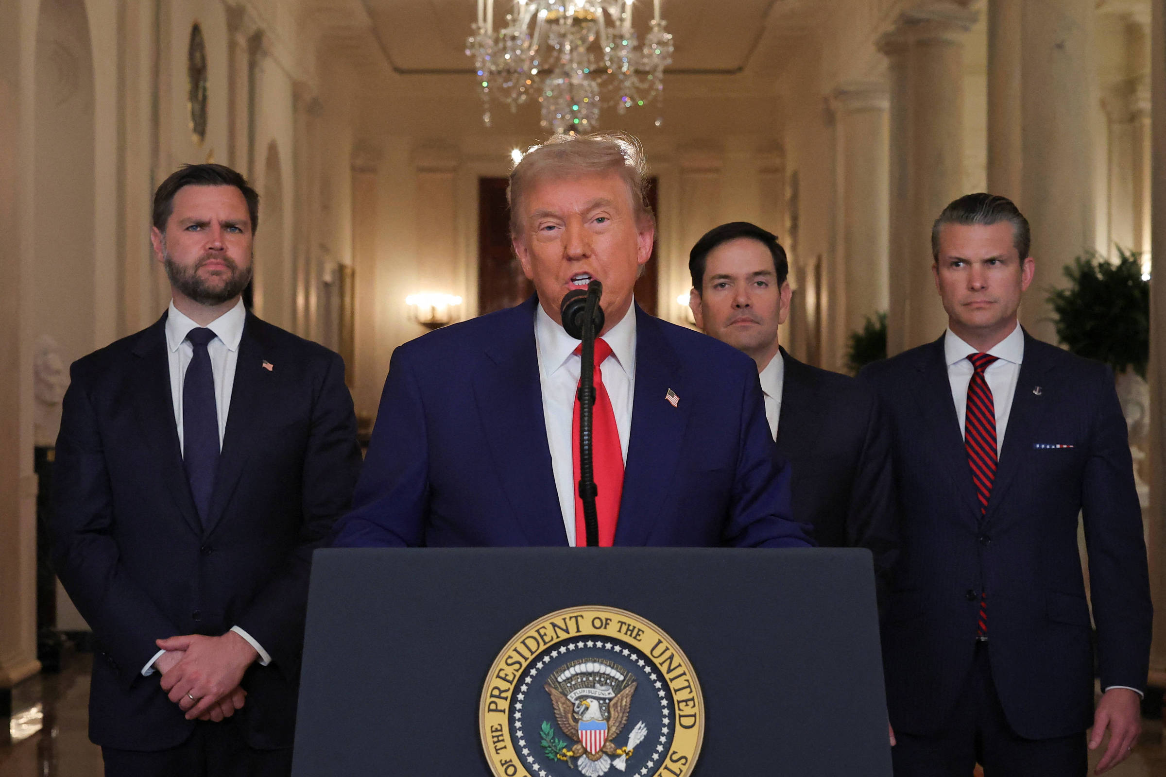 A imagem mostra um homem em um terno azul com uma gravata vermelha, falando ao microfone em um evento oficial. Ele está posicionado em frente a um púlpito com o selo da presidência dos Estados Unidos. Atrás dele, há três homens em trajes formais, um deles à esquerda com um terno escuro e gravata, e os outros dois à direita, um com um terno claro e o outro com um terno escuro e gravata listrada. O ambiente é um salão com lustres e decoração clássica.