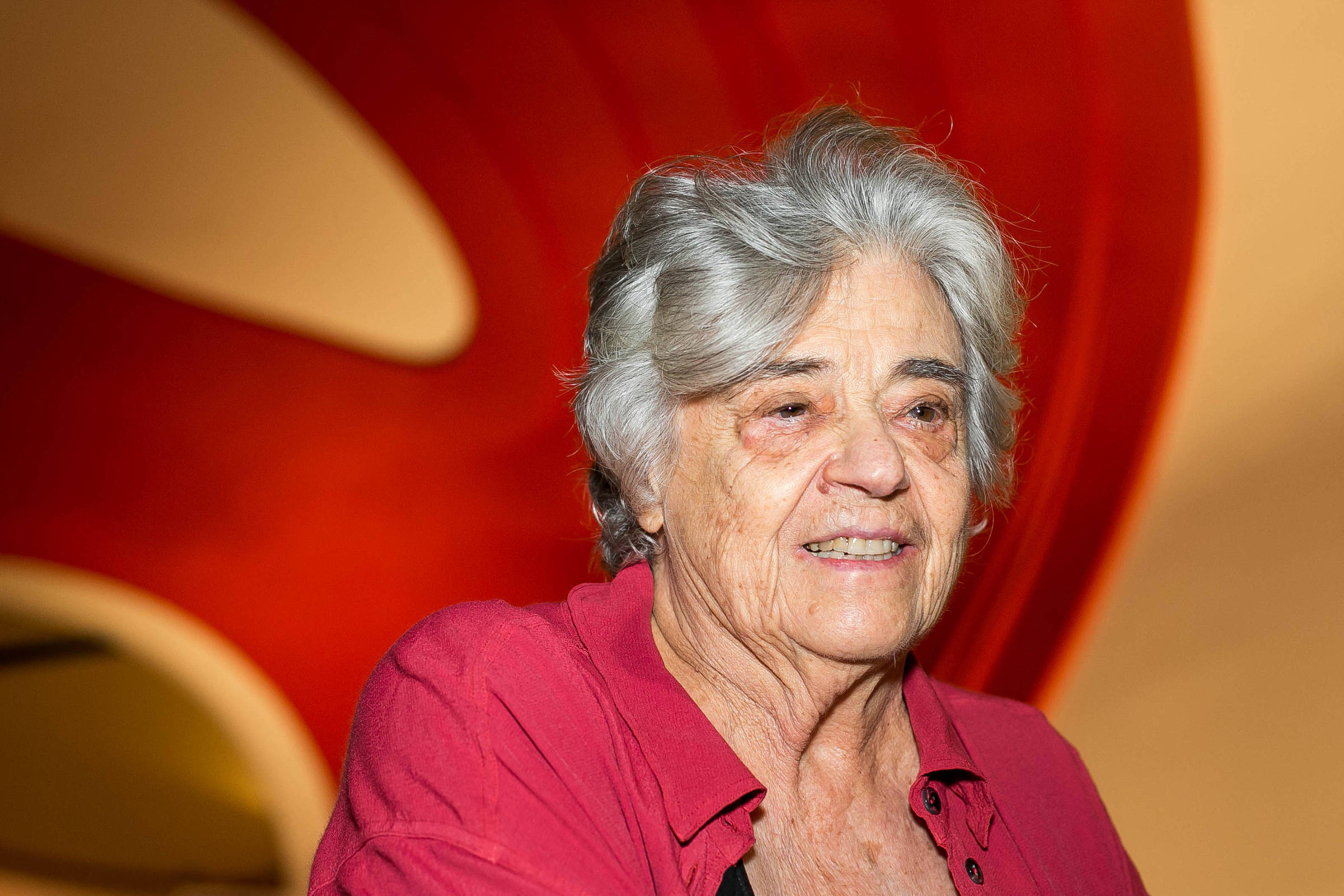 A imagem mostra uma mulher idosa com cabelo grisalho e uma blusa vermelha. Ela está sorrindo e parece estar em um ambiente com um fundo colorido, onde predominam tons de vermelho e laranja. A iluminação é suave, destacando seu rosto e expressões.
