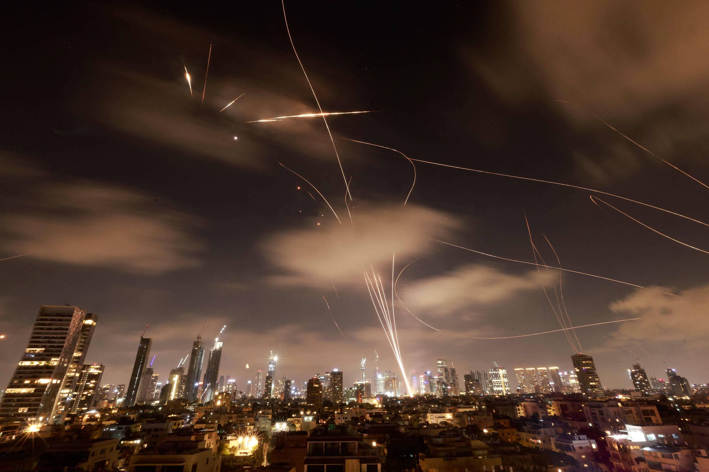 A imagem mostra uma vista noturna de uma cidade, onde arranha-céus iluminados se destacam contra o céu escuro. No céu, há várias trajetórias de luz, possivelmente de foguetes ou mísseis, que criam um efeito visual dinâmico. As nuvens estão presentes, e a cena transmite uma sensação de atividade intensa.