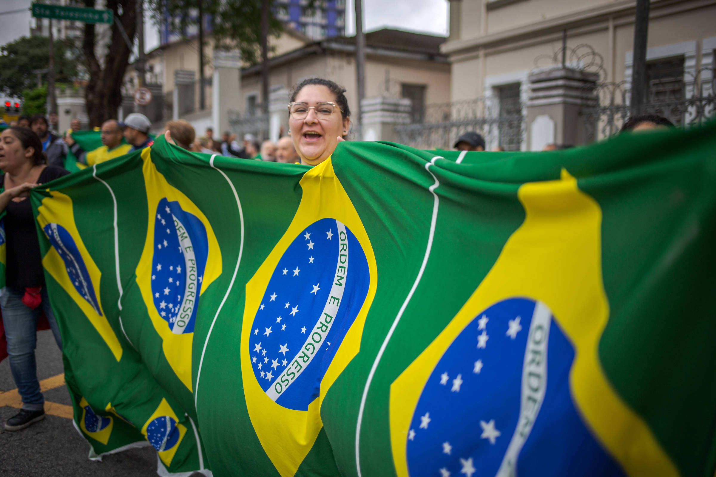 A imagem mostra um grupo de pessoas participando de uma manifestação, segurando uma grande bandeira do Brasil. A bandeira é predominantemente verde e amarelo, com um grande losango azul no centro, que contém estrelas brancas. Uma mulher em destaque, com óculos e cabelo preso, sorri enquanto segura a bandeira. Ao fundo, é possível ver outras pessoas e um ambiente urbano.
