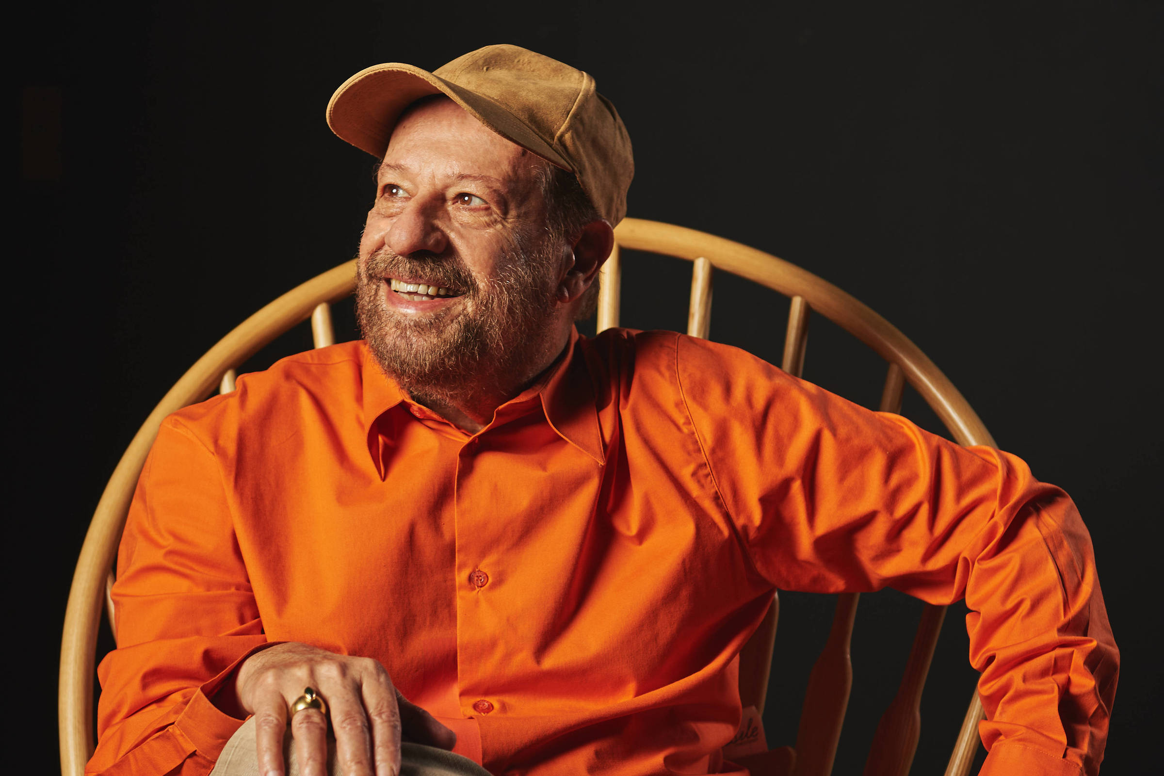 Um homem está sentado em uma cadeira de madeira com um design de palhinha. Ele usa uma camisa laranja e um boné claro. O homem tem uma barba e está sorrindo, com uma perna cruzada sobre a outra. O fundo da imagem é escuro, destacando a figura do homem e a cadeira.