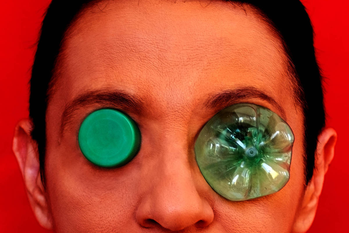 A imagem apresenta um rosto humano com um fundo vermelho. O rosto tem um cabelo curto e liso, e os olhos estão cobertos por dois objetos: um disco verde e uma garrafa plástica verde. A expressão facial é neutra, e a composição é bastante contrastante devido ao fundo e aos objetos utilizados.