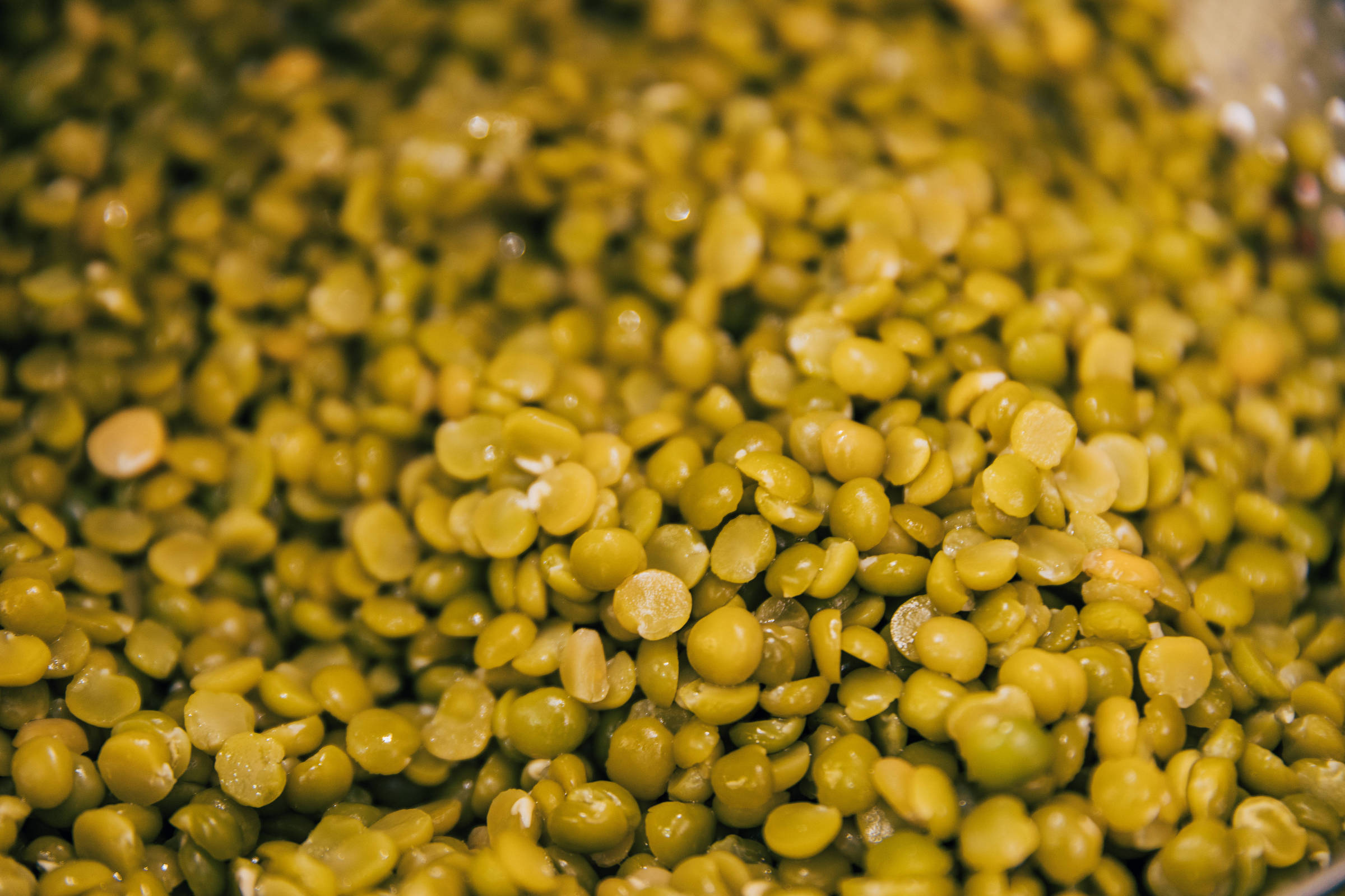 A imagem mostra uma grande quantidade de grãos de ervilha verde, dispostos de forma desordenada. Os grãos são pequenos, arredondados e apresentam uma coloração verde vibrante, com algumas variações de tonalidade. A superfície dos grãos parece brilhante, indicando que estão frescos ou foram cozidos recentemente.