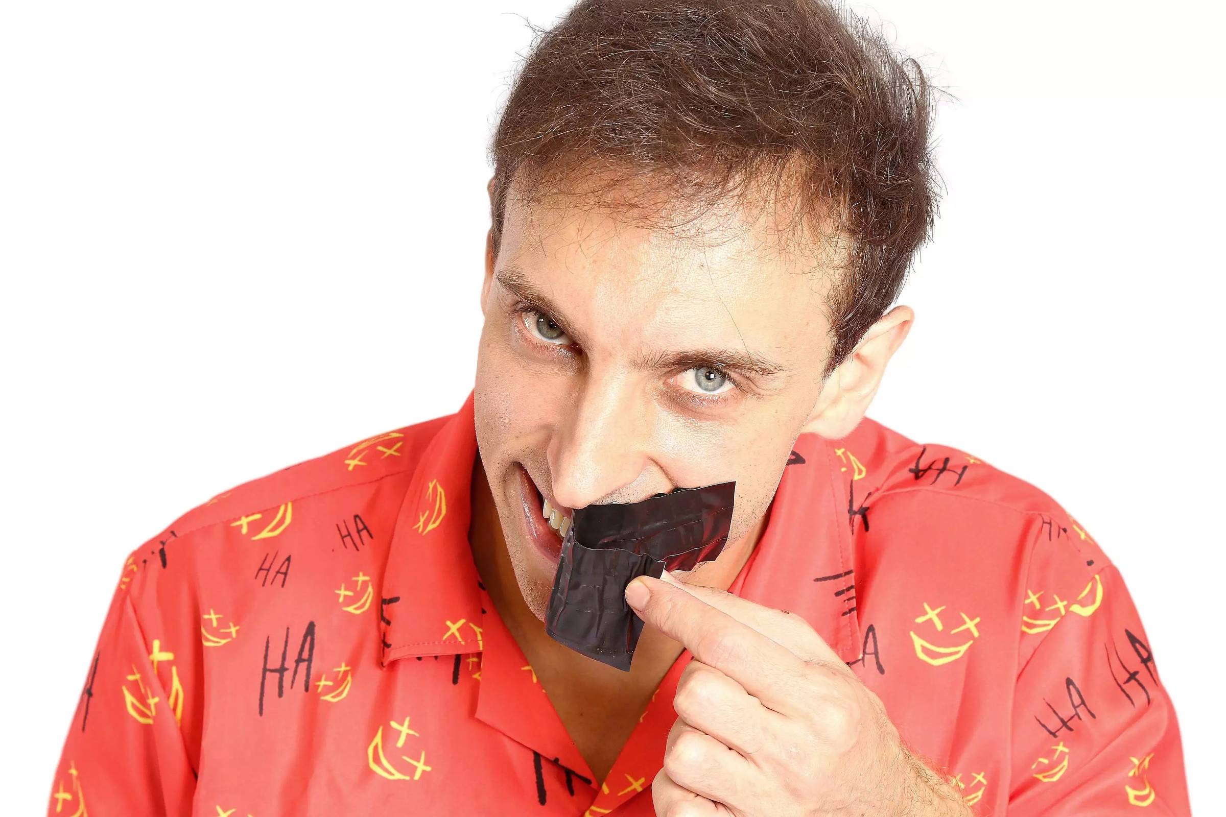 Um homem está segurando um pedaço de fita preta perto da boca, enquanto olha para a câmera. Ele usa uma camisa vermelha com estampas de risadas e símbolos. O fundo é branco, destacando a figura do homem.
