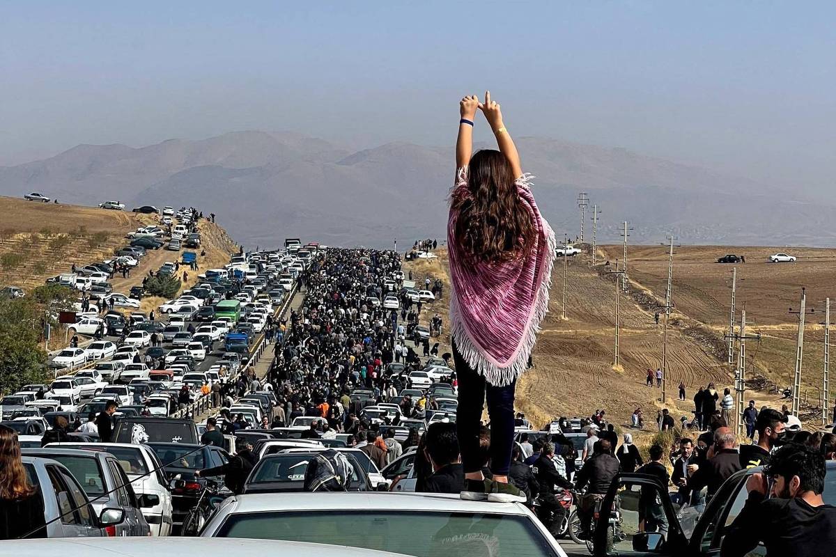 A imagem mostra uma grande multidão de pessoas e veículos parados em uma estrada. No primeiro plano, uma jovem está em cima de um carro, com as mãos levantadas em sinal de protesto. Ao fundo, é possível ver uma longa fila de carros e uma multidão de pessoas caminhando. A paisagem é montanhosa e o céu está claro.