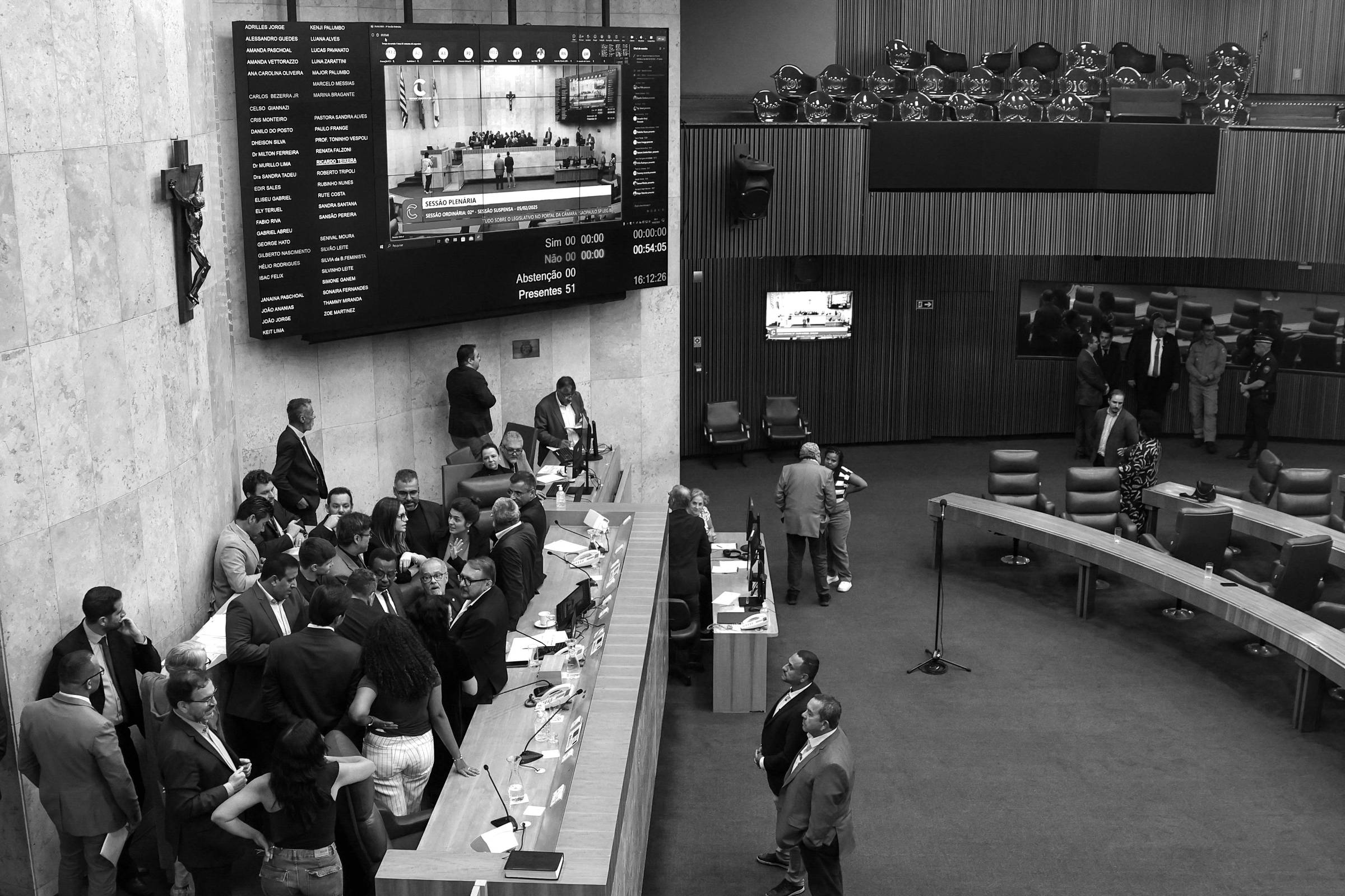 A imagem mostra o interior de um plenário legislativo, onde várias pessoas estão reunidas em torno de uma mesa em formato de U. No fundo, há um grande painel eletrônico exibindo informações sobre a sessão. Algumas pessoas estão de pé, enquanto outras estão sentadas. O ambiente é bem iluminado e possui paredes de tom claro, com cadeiras azuis dispostas em fileiras. Um crucifixo é visível na parede à esquerda.