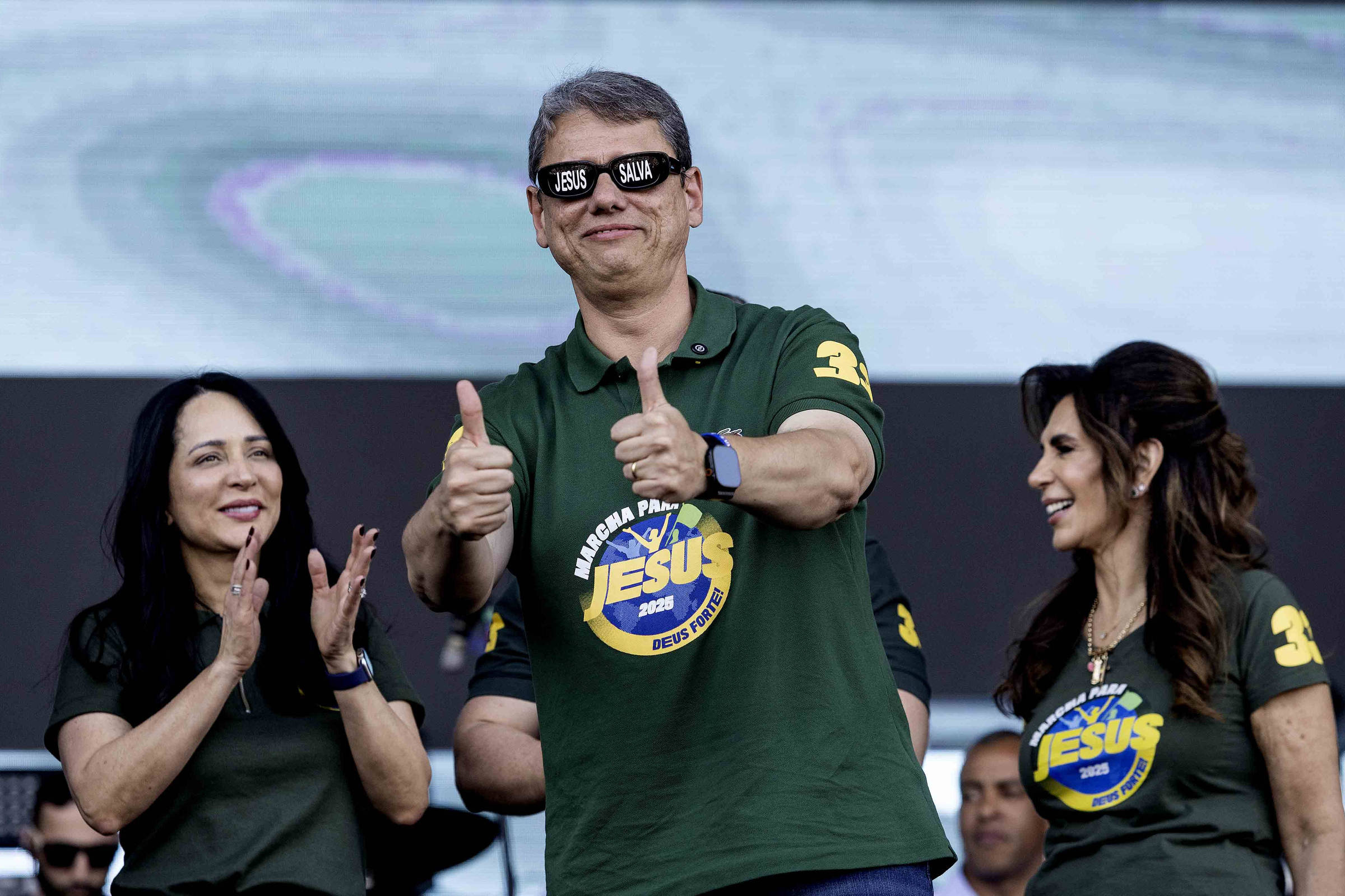 A imagem mostra três pessoas em um palco durante um evento. O homem no centro, usando óculos escuros, faz um sinal de positivo com as duas mãos e está vestido com uma camiseta verde com um logo em destaque. À sua esquerda, uma mulher aplaude, enquanto à direita, outra mulher sorri. Todos parecem estar se divertindo em um ambiente festivo.