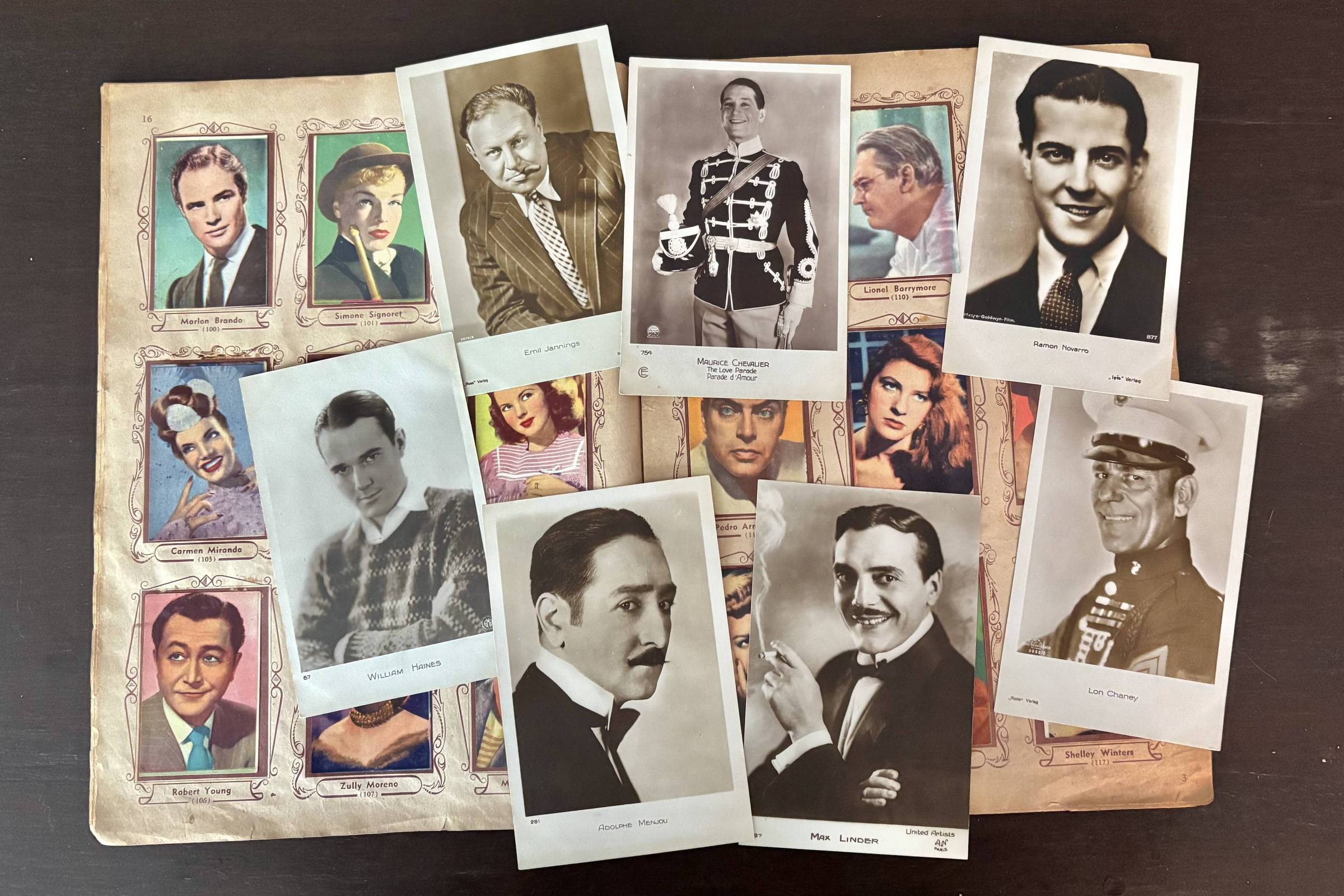 A imagem mostra uma coleção de fotografias de celebridades, dispostas sobre uma superfície escura. As fotos incluem retratos de atores e atrizes, como Marlon Brando, Carmen Miranda, e Maurice Chevalier, entre outros. As imagens estão organizadas em uma página decorada, com bordas ornamentadas, e algumas fotos estão sobrepostas. Cada retrato possui uma legenda com o nome do indivíduo e, em alguns casos, o ano de nascimento. A composição é rica em detalhes e apresenta uma variedade de estilos e poses.