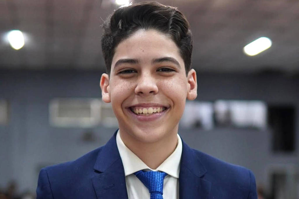 Um jovem sorridente está posando para a foto em um ambiente interno. Ele usa um terno escuro e uma gravata azul, com uma camisa branca por baixo. O fundo é desfocado, mas parece haver várias pessoas e mesas ao redor, sugerindo um evento social ou celebração.