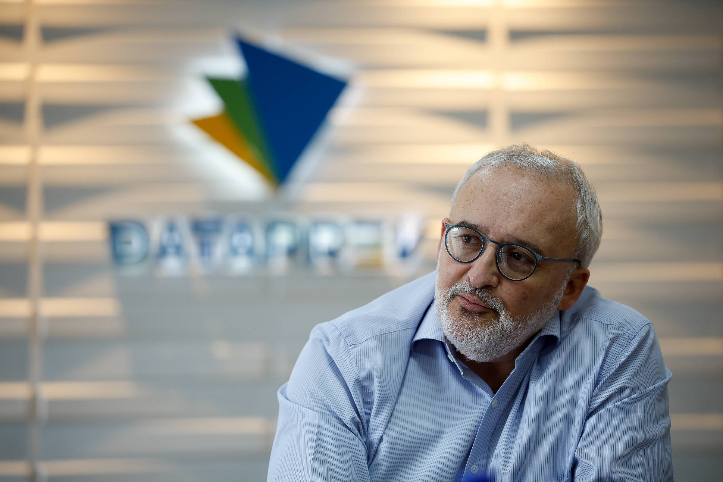 Um homem com cabelo grisalho e barba, usando óculos, está sentado em um ambiente de escritório. Ele veste uma camisa azul clara e parece estar em uma conversa ou reflexão. Ao fundo, há um logotipo colorido que diz