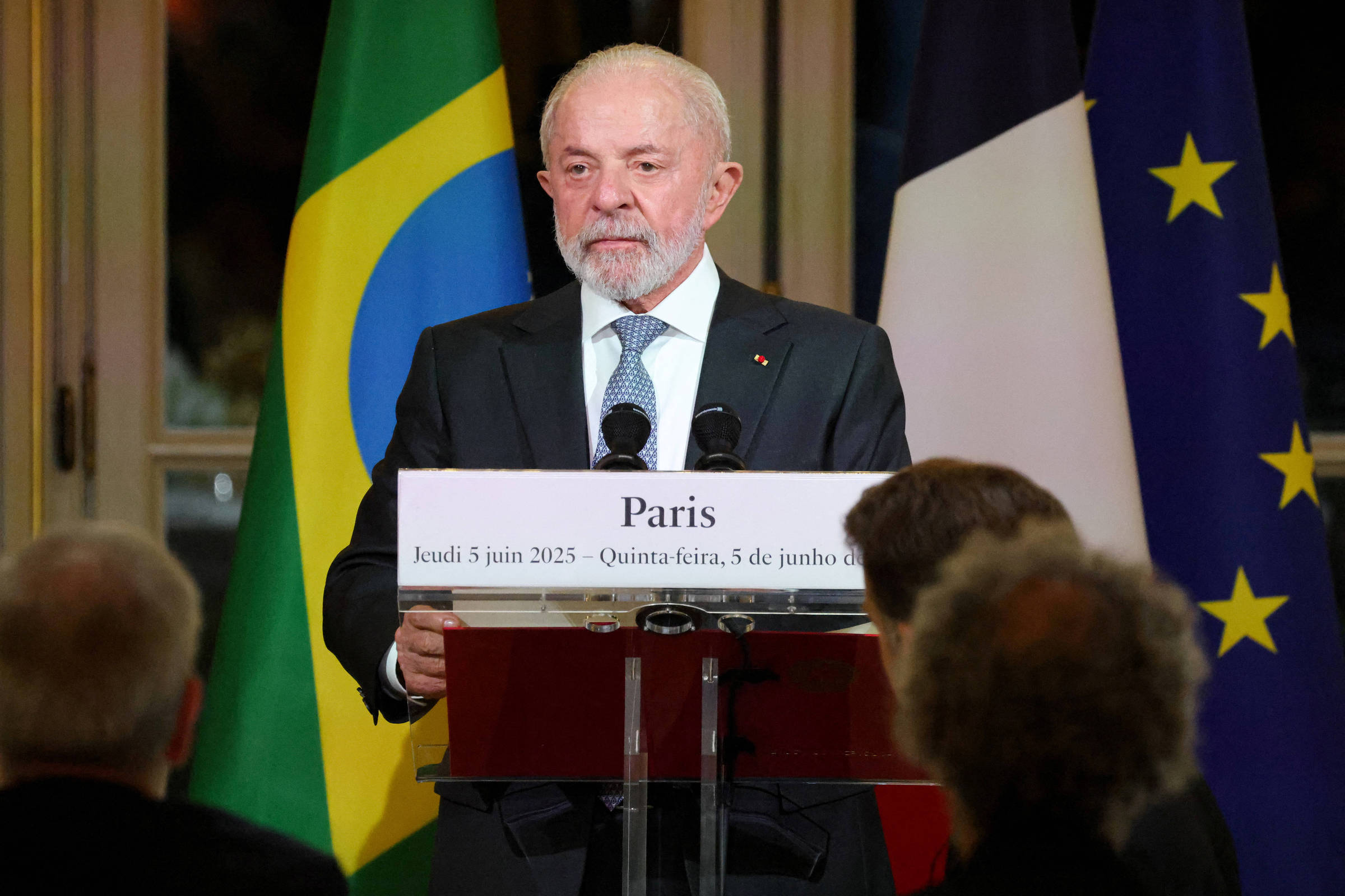 Um homem de cabelo grisalho e barba, vestido com um terno escuro e gravata, está em pé atrás de um púlpito de vidro. Ele parece estar fazendo um discurso. Ao fundo, há bandeiras do Brasil, da França e da União Europeia. Uma placa na frente dele diz