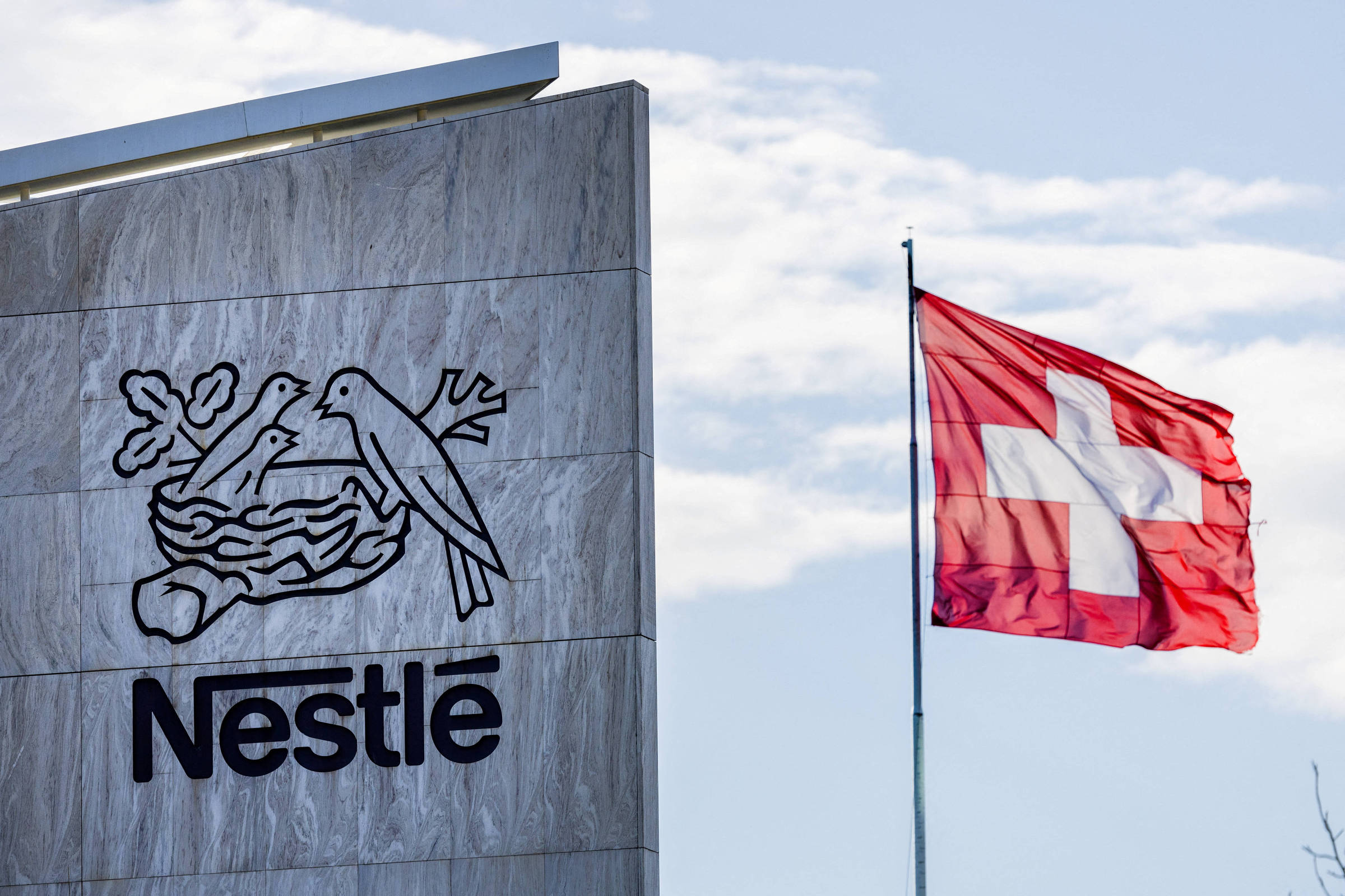 A imagem mostra o logotipo da Nestlé em uma placa de pedra, com um desenho de um ninho e pássaros. Ao lado, está a bandeira da Suíça, que é vermelha com uma cruz branca no centro. O céu está claro e há algumas nuvens.