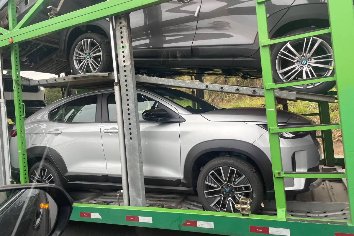 A imagem mostra um caminhão transportando carros. Na parte inferior, há um carro SUV de cor prata, enquanto na parte superior, um carro de cor cinza está visível. Ambos os veículos parecem ser modelos da marca BMW, com detalhes visíveis nas rodas e na carroceria. O caminhão é de cor verde e possui uma estrutura metálica que suporta os carros.
