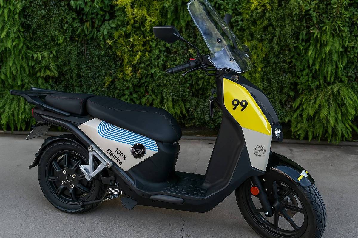 Uma scooter elétrica de cor preta e branca, com detalhes em amarelo. O número 99 está visível na parte frontal. A scooter possui um design moderno e é estacionada em uma superfície clara, com um fundo de plantas verdes ao fundo.