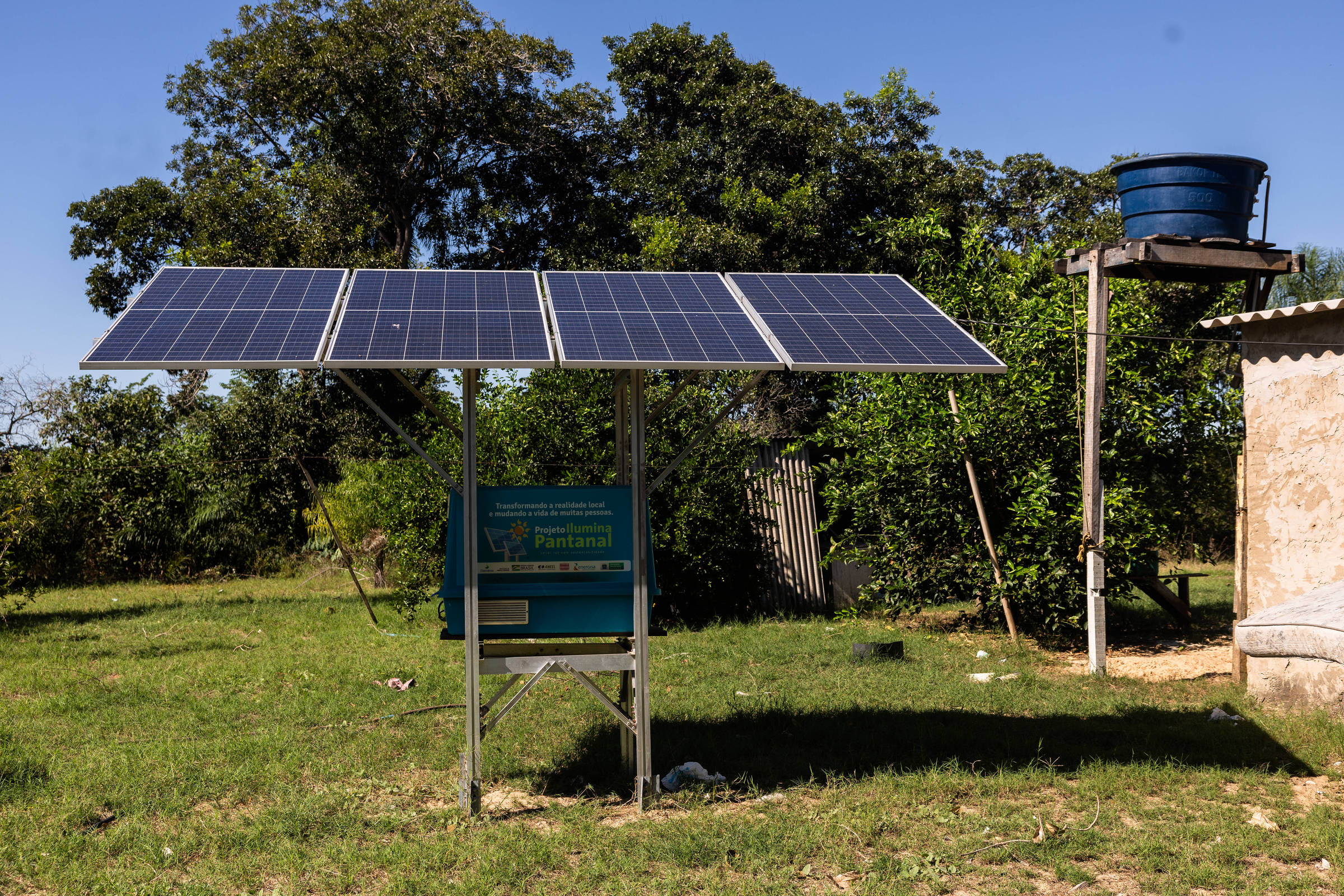 A imagem mostra um sistema de energia solar instalado em uma área rural. Há um conjunto de painéis solares montados em uma estrutura metálica, com um fundo de vegetação e árvores. Ao lado, há um tanque de água elevado. O céu está limpo e ensolarado, e a grama é verde.