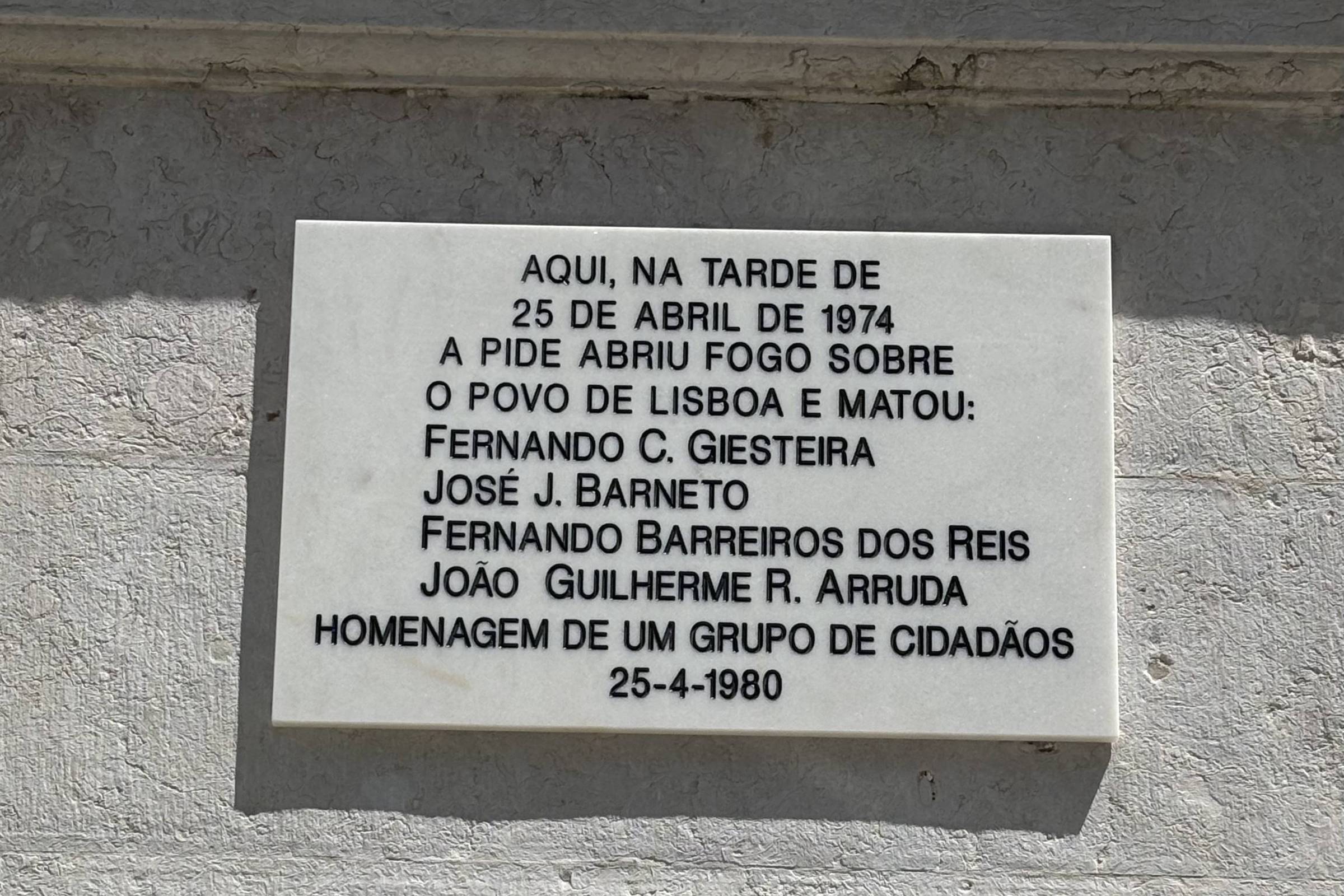 Uma placa de mármore branca fixada em uma parede, com texto em letras pretas. O texto menciona um evento ocorrido na tarde de 25 de abril de 1974, quando a PIDE abriu fogo sobre o povo de Lisboa, resultando na morte de quatro pessoas: Fernando C. Giesteira, José J. Barneto, Fernando Barreiros dos Reis e João Guilherme R. Arruda. A placa é uma homenagem de um grupo de cidadãos e foi instalada em 25 de abril de 1980.