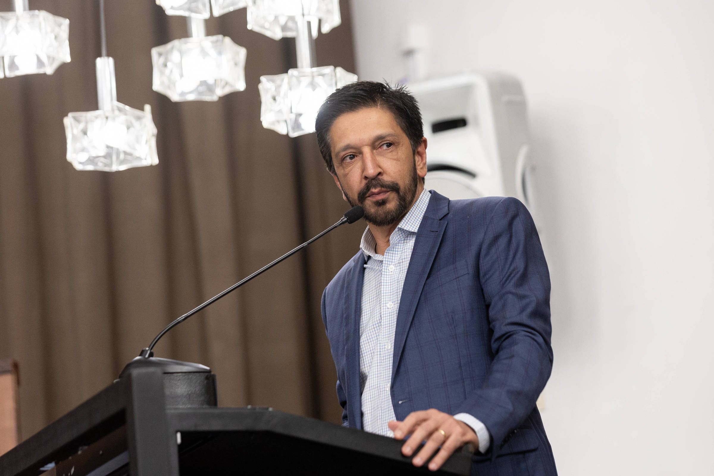 Um homem está em pé atrás de um púlpito, falando ao microfone. Ele usa um terno azul escuro e uma camisa clara. Ao fundo, há cortinas marrons e um lustre com luminárias de cristal. O ambiente parece ser um salão de eventos.