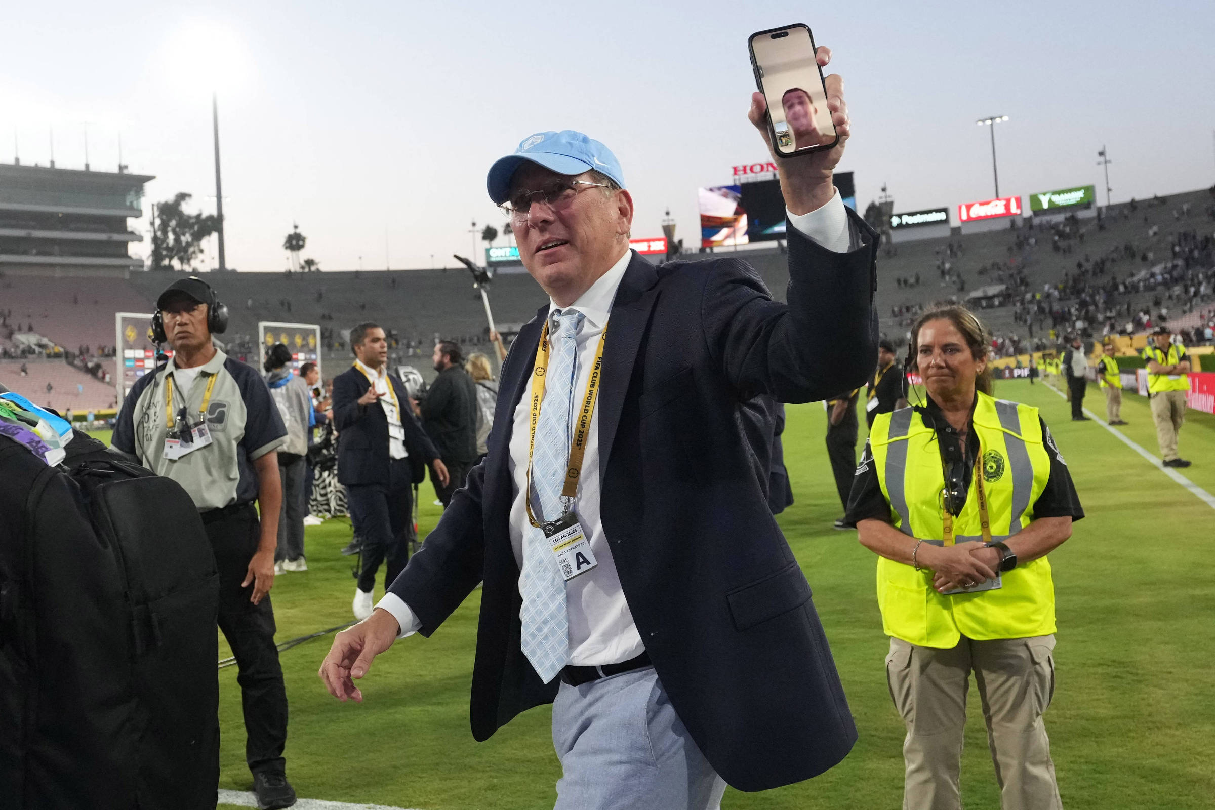 Um homem vestido com um terno escuro e uma gravata clara, usando um boné azul, está caminhando pelo campo de futebol. Ele segura um celular na mão, mostrando uma videochamada. Ao fundo, há pessoas e um estádio parcialmente cheio. Uma mulher com colete amarelo está ao lado dele, observando.