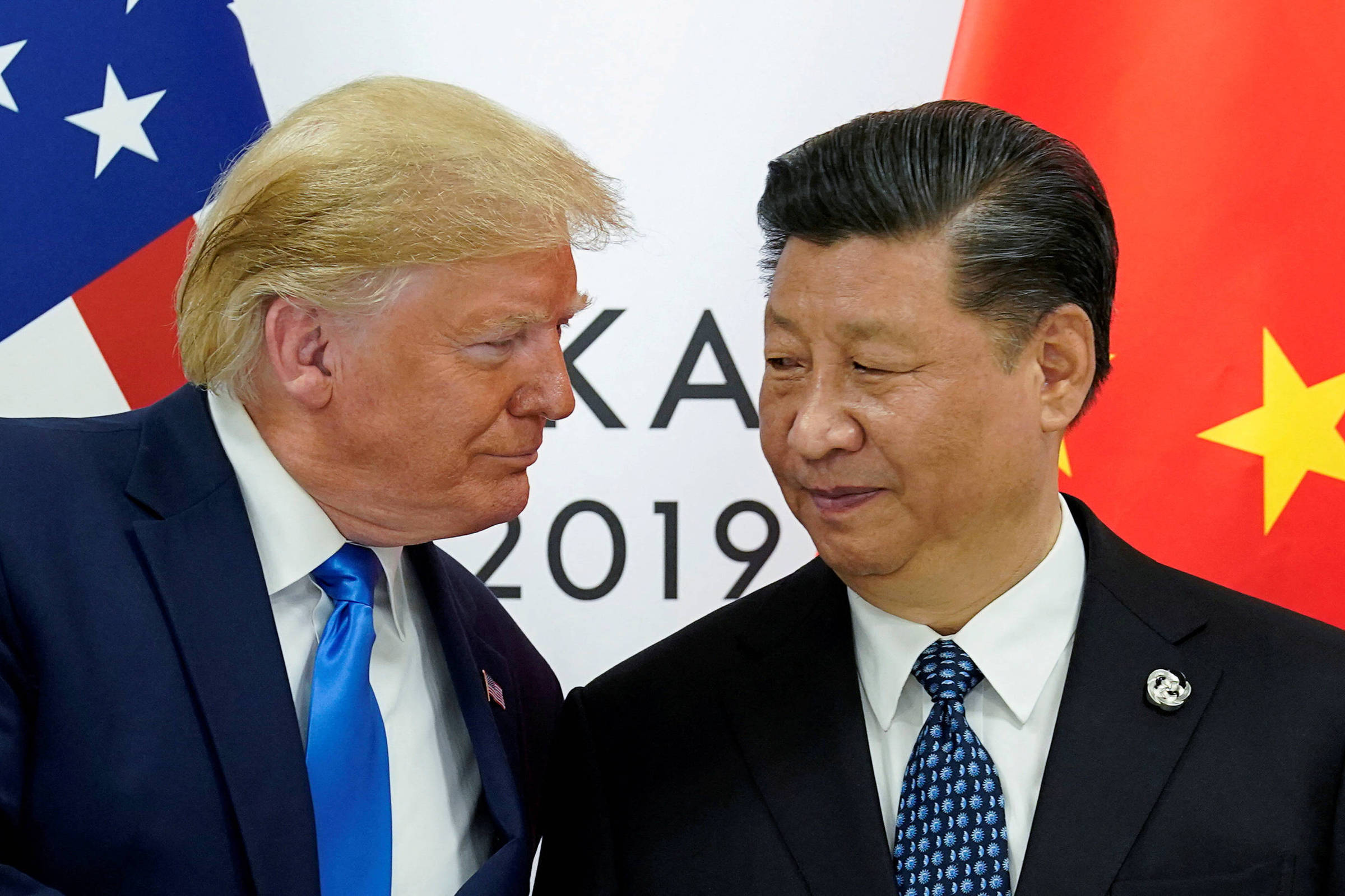 A imagem mostra dois homens em um encontro, um deles com cabelo loiro e terno escuro, inclinando-se em direção ao outro, que tem cabelo escuro e usa um terno preto. Ao fundo, há bandeiras dos Estados Unidos e da China. A expressão facial de ambos sugere um momento de interação amigável.