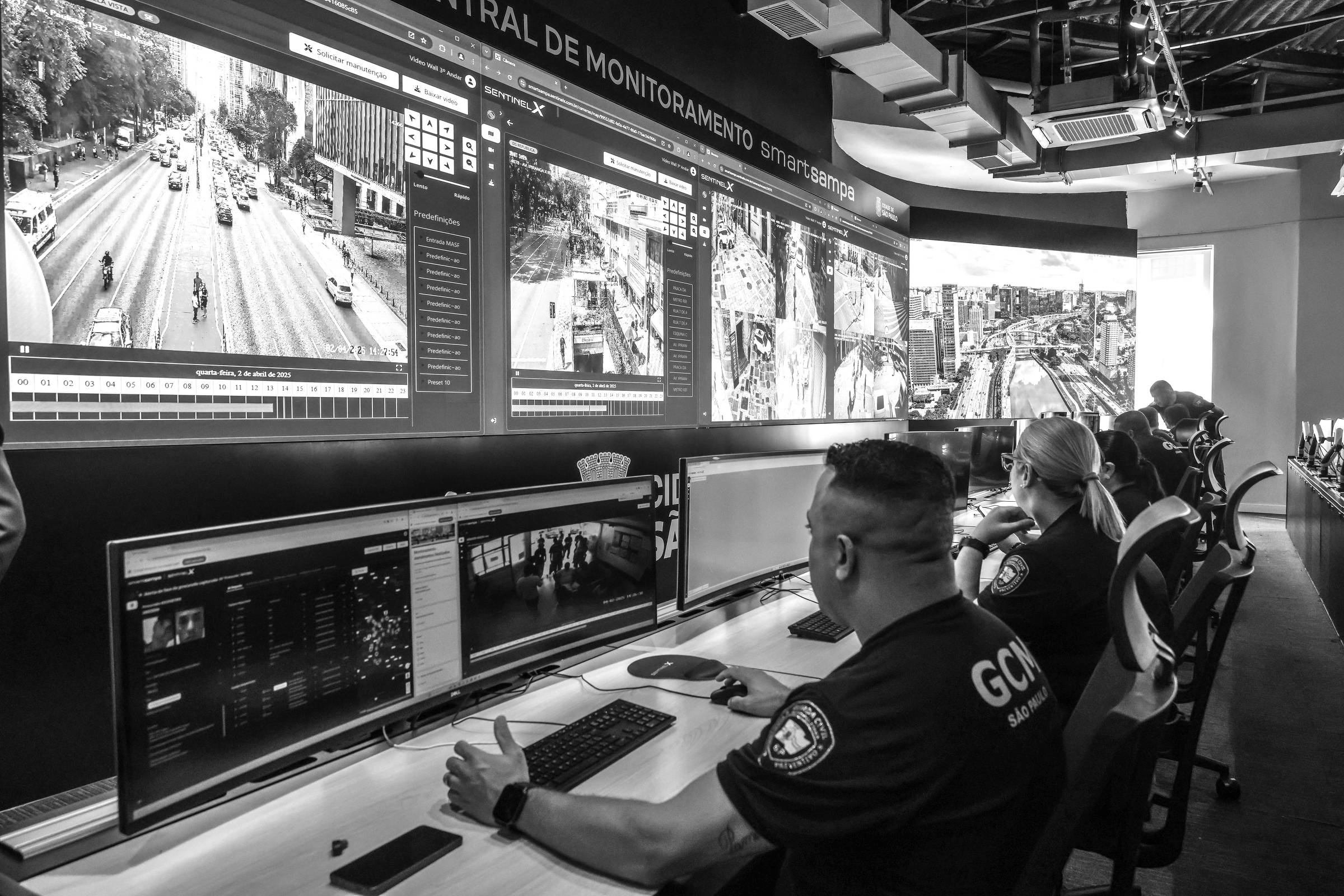 A imagem mostra uma central de monitoramento com várias telas exibindo imagens de tráfego e segurança. Dois operadores estão sentados em frente a computadores, observando as imagens. As telas mostram diferentes ângulos de uma área urbana, com veículos e pedestres visíveis. O ambiente é moderno e bem iluminado, com um design funcional.