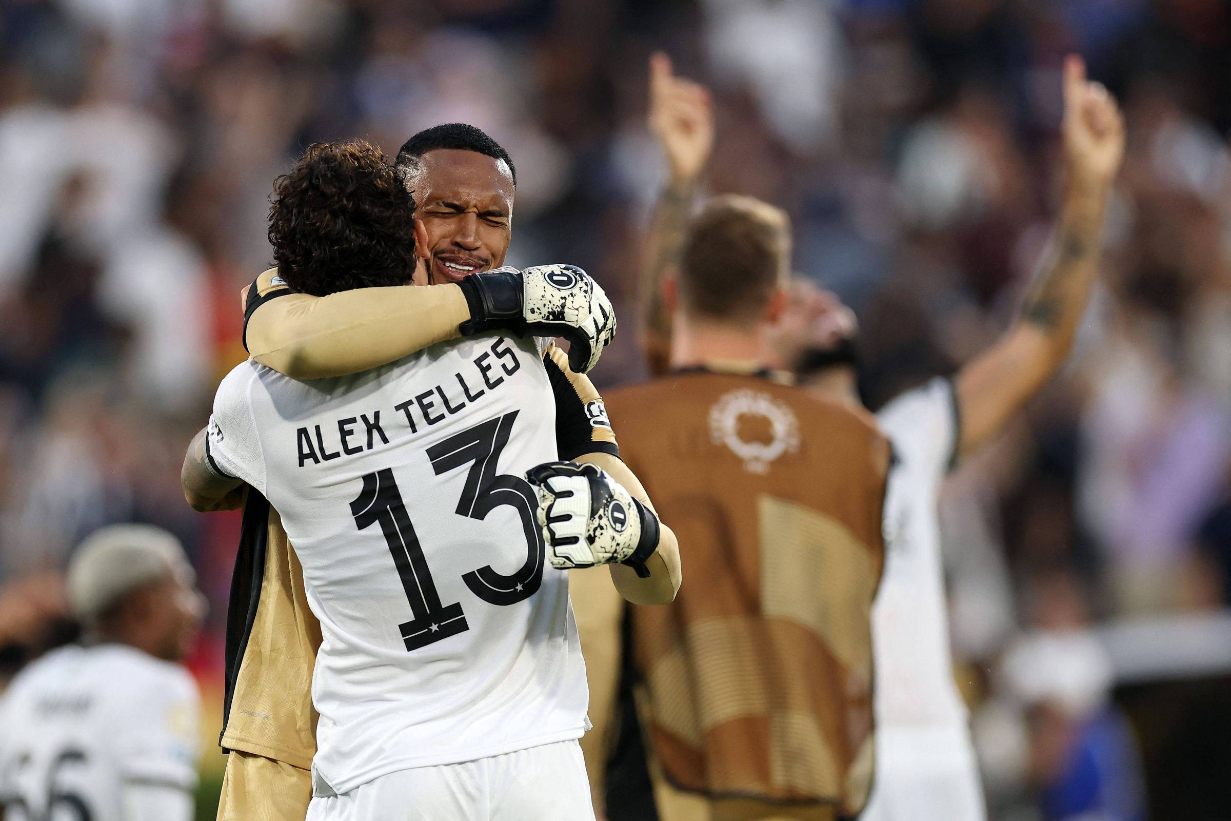 Na imagem, dois jogadores de futebol estão se abraçando em um momento de celebração. Um deles está usando uma camisa branca com o nome