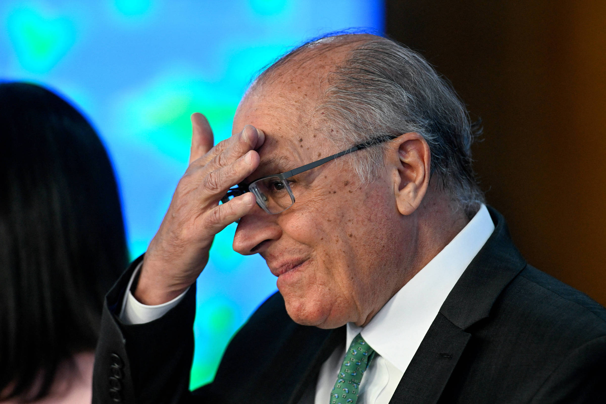 Um homem de cabelos grisalhos e óculos, vestindo um terno escuro e uma gravata verde, está em um evento. Ele está com a mão na testa, como se estivesse pensativo ou preocupado. Ao fundo, há uma tela com um padrão colorido, que parece ser uma apresentação. Uma mulher com cabelo longo e escuro está parcialmente visível à esquerda.