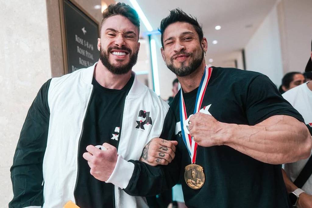 Dois homens posam juntos em um ambiente interno. Um deles usa uma jaqueta branca e preta e segura uma pasta amarela, enquanto o outro está vestido com uma camiseta preta e exibe uma medalha de ouro ao redor do pescoço. Ambos estão sorrindo e fazendo um gesto de força com os braços.