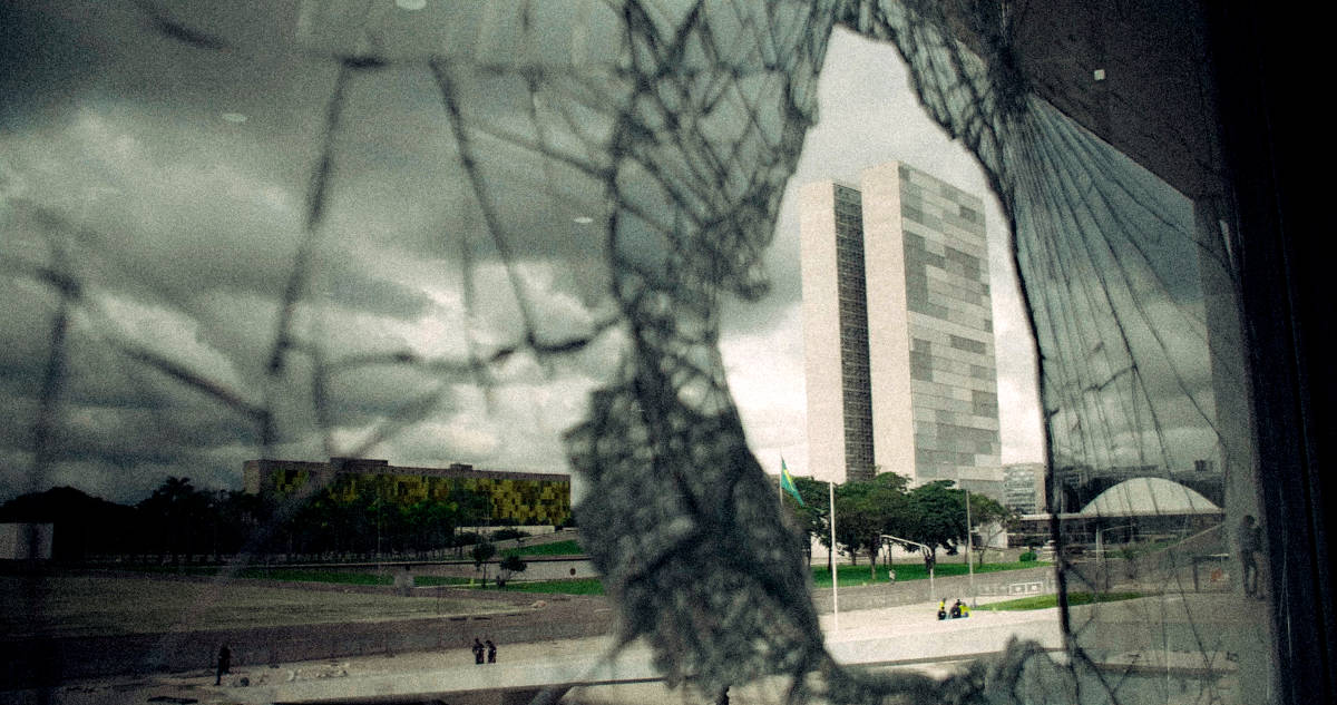 A imagem mostra uma vista de um edifício moderno através de um vidro quebrado. O vidro apresenta uma grande fissura, e ao fundo é possível ver um céu nublado com nuvens escuras. O edifício à esquerda tem uma fachada verde e o edifício à direita é mais alto e possui uma fachada em tons de cinza. No chão, há algumas pessoas e bandeiras visíveis.