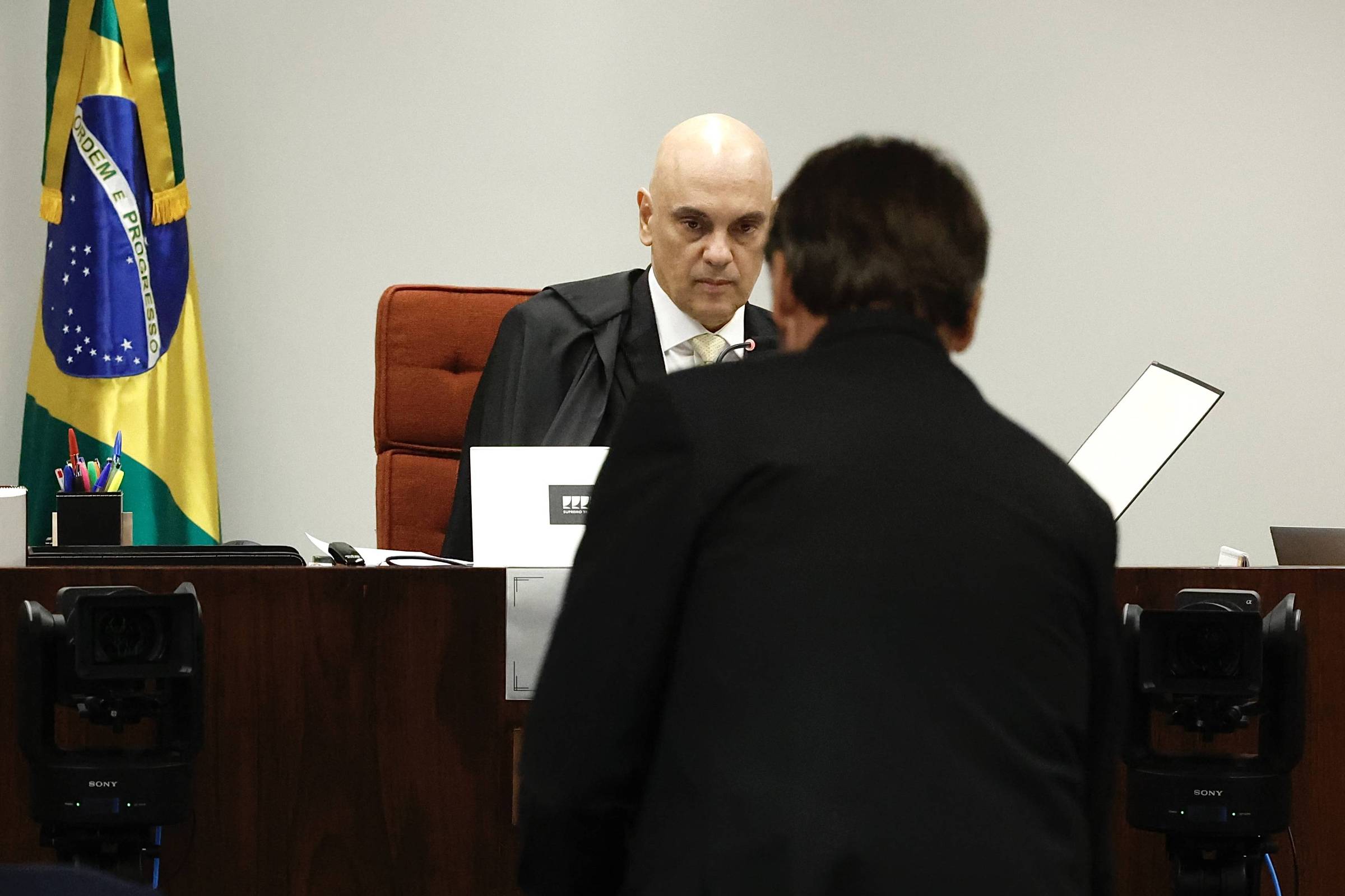 A imagem mostra um juiz com cabelo calvo e vestindo uma toga, sentado em uma mesa de tribunal. Ele está olhando para um homem de costas, que está de pé em frente a ele. Ao fundo, há uma bandeira do Brasil e equipamentos de gravação. O ambiente é de um tribunal, com uma atmosfera formal.